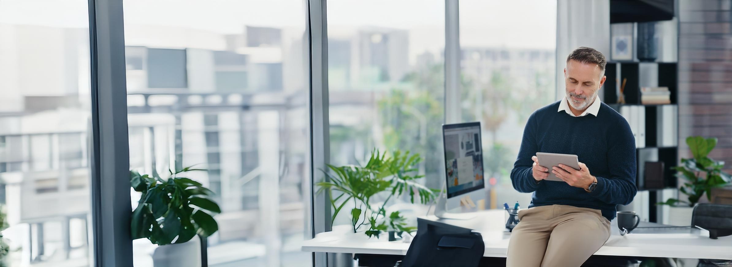 A confident business professional sitting on a desk in a modern office space, reviewing a tablet. The bright, open space features large windows, greenery, and contemporary office decor, creating an atmosphere of productivity and innovation.