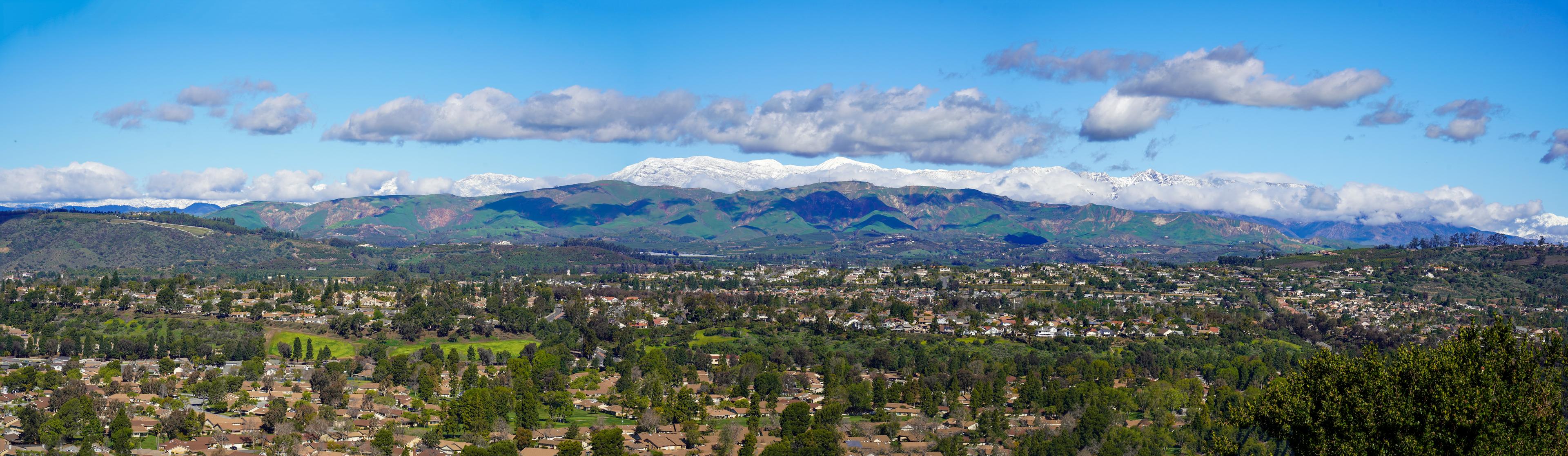 Panoramic view of a residential area in Santa Rosa, California, surrounded by lush green hills and distant snow-capped mountains under a bright blue sky with scattered clouds. 