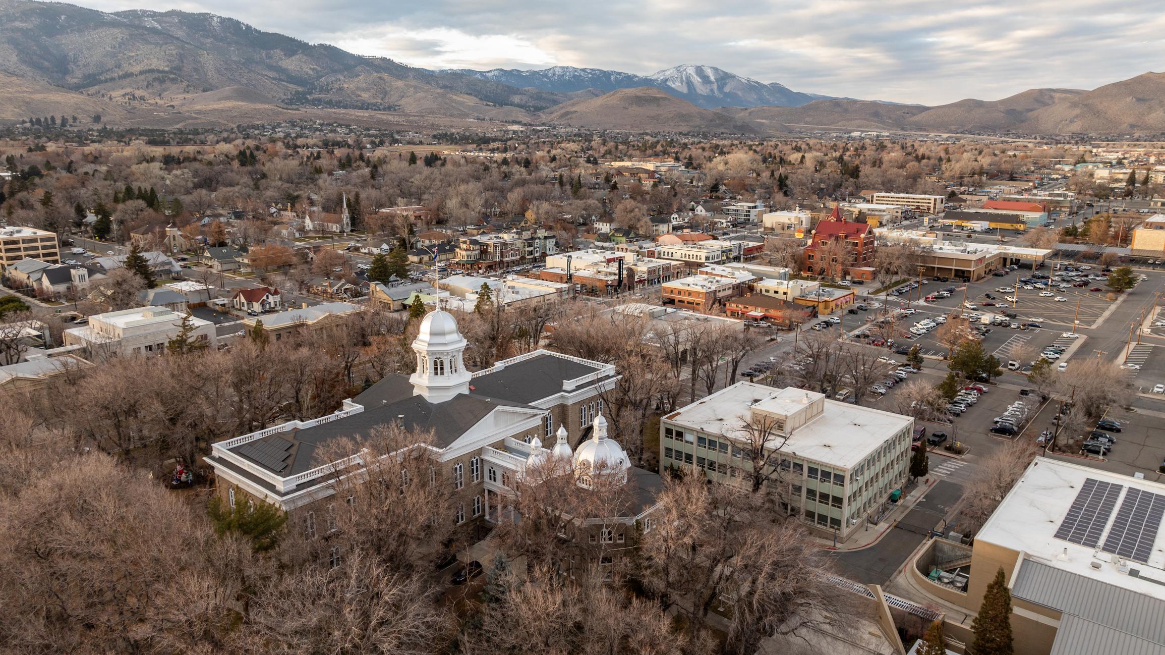 A view of downtown Carson City, Nevada, showcasing the Nevada State Capitol building among tree-lined streets and small buildings. The surrounding city and distant snow-dusted mountains create a scenic backdrop under a cloudy sky.