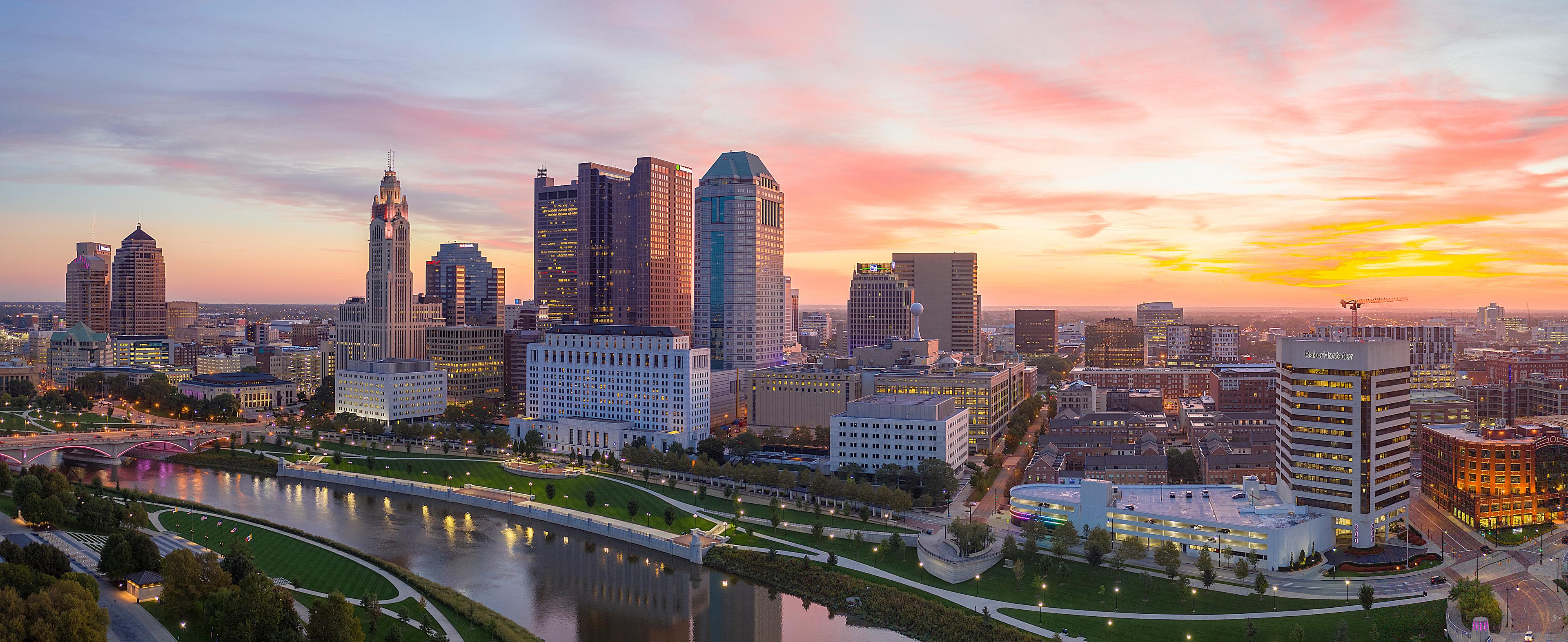 Panoramic sunset view of downtown Columbus, Ohio, with the Scioto River in the foreground and the city skyline glowing under a vibrant, colorful sky.