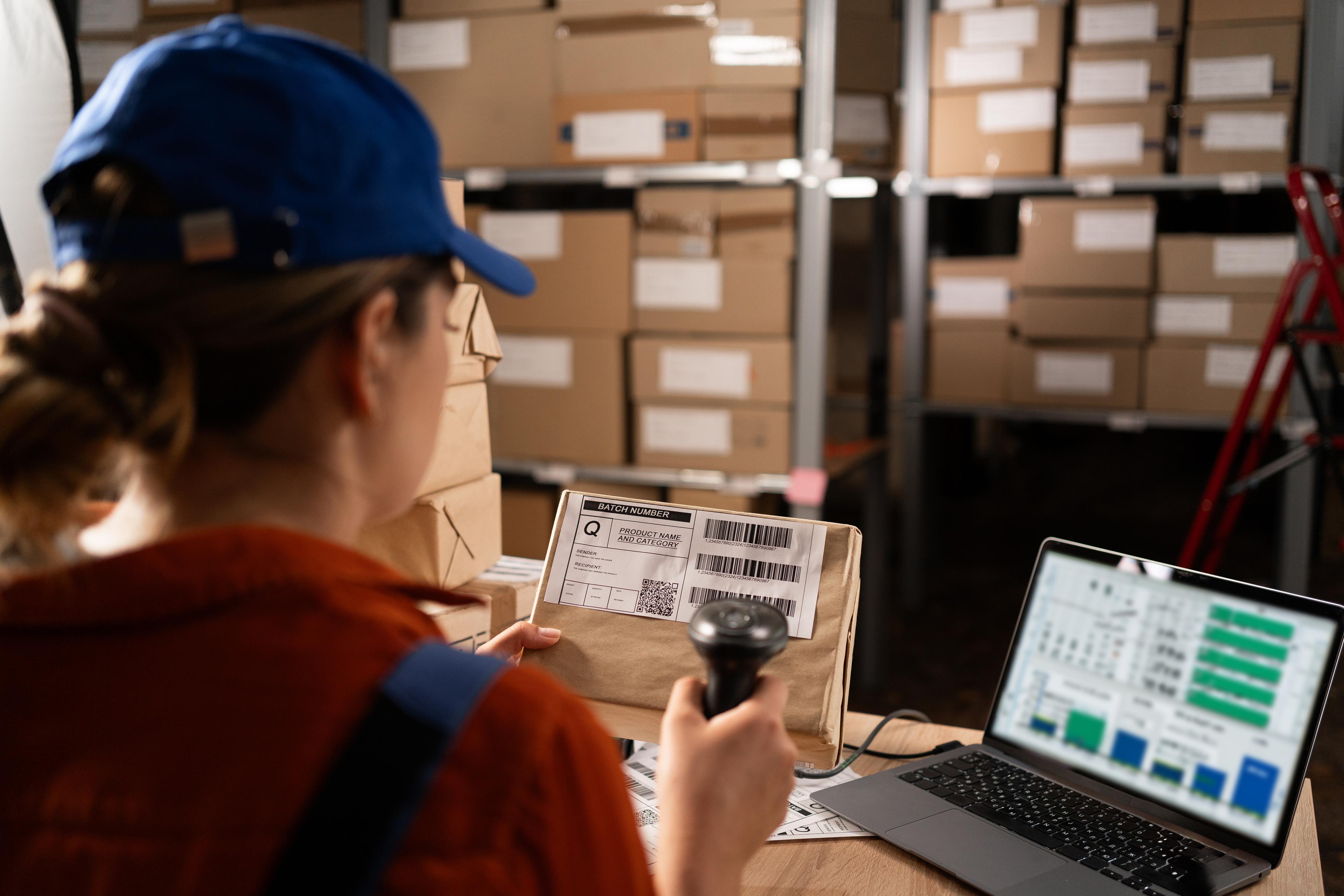 A worker in a warehouse scanning a package with a barcode scanner. The person is sitting at a desk with a laptop displaying inventory management software. Shelves filled with labeled boxes are visible in the background, indicating a logistics or inventory control process.