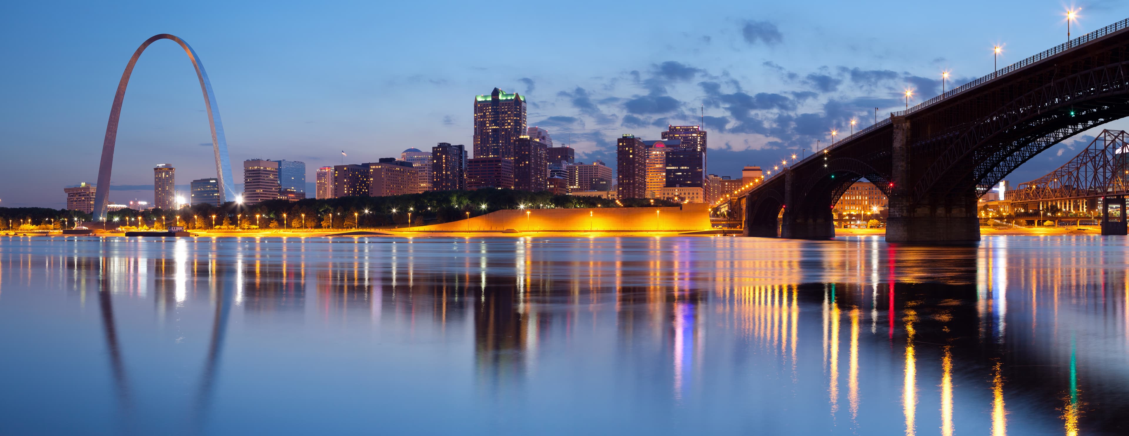 St. Louis skyline at twilight with the Gateway Arch and Eads Bridge reflected in the calm waters of the Mississippi River, city lights glowing against a dusky blue sky.