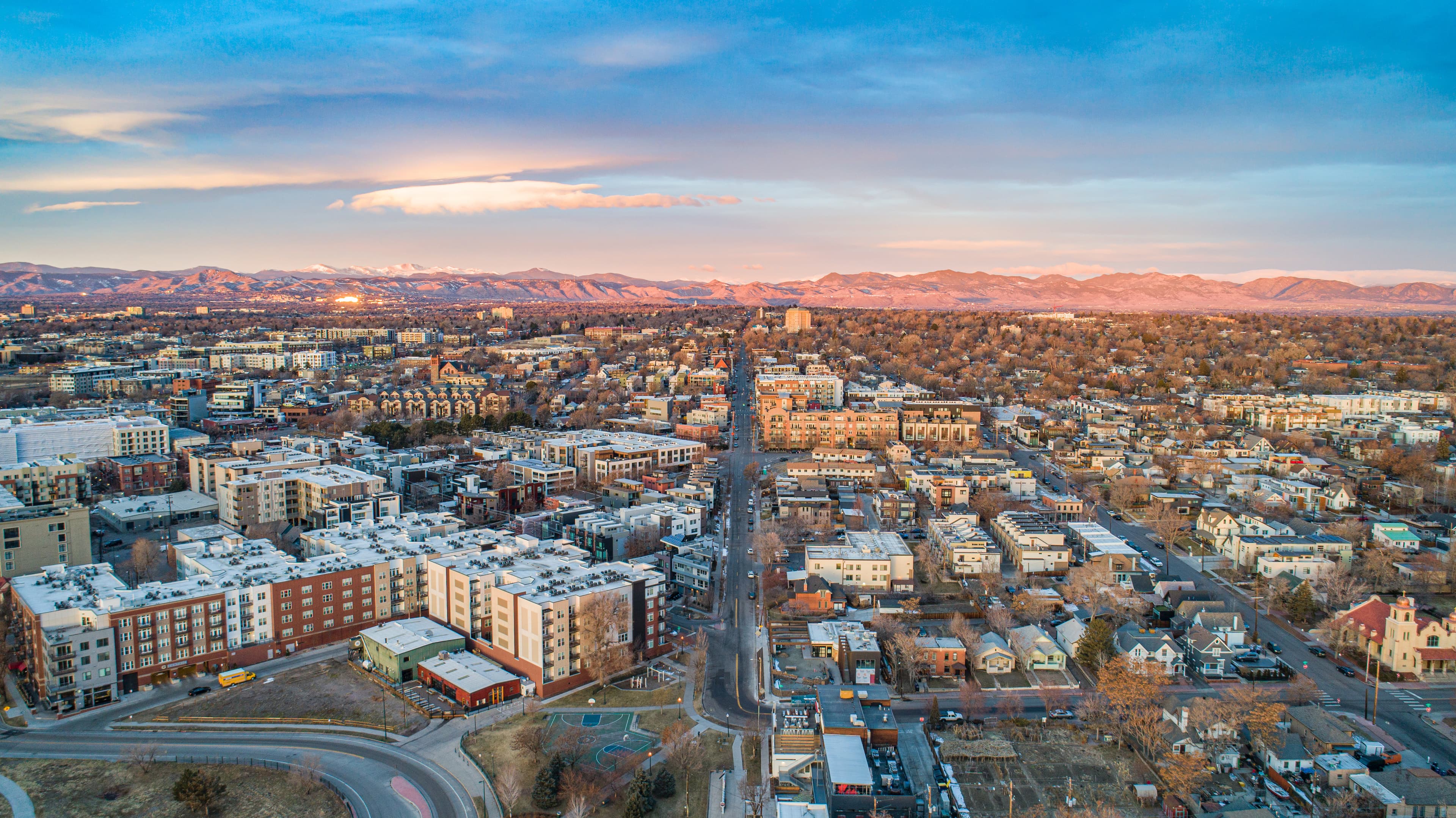 Aerial view of Fort Collins, Colorado at sunset, showcasing a mix of residential and commercial buildings, tree-lined streets, and the distant Rocky Mountains bathed in warm light under a pastel-colored sky.
