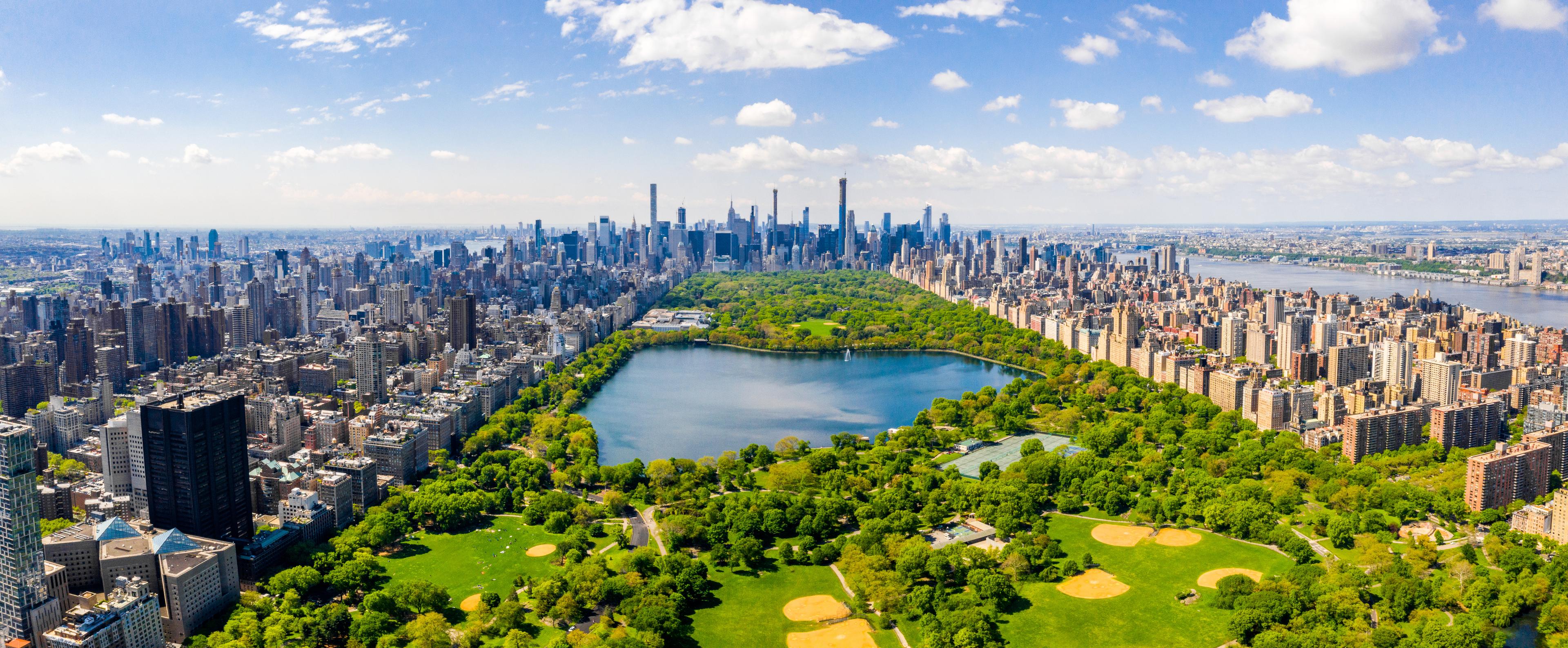 An aerial view of New York City's Central Park, surrounded by densely packed skyscrapers and urban buildings, with the Hudson River visible on the right under a bright blue sky with scattered clouds.