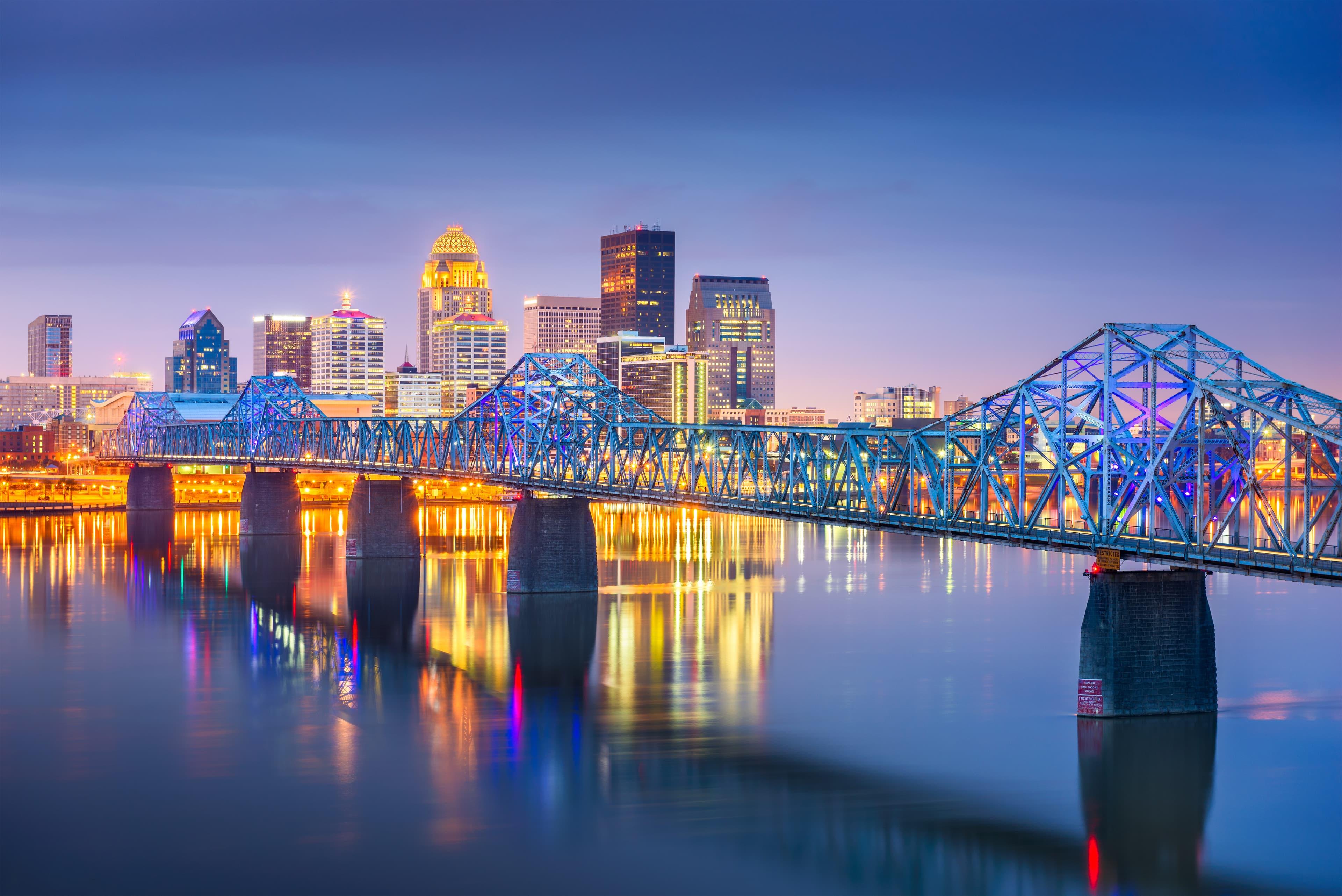 Louisville skyline at dusk with illuminated bridge reflecting over the Ohio River.