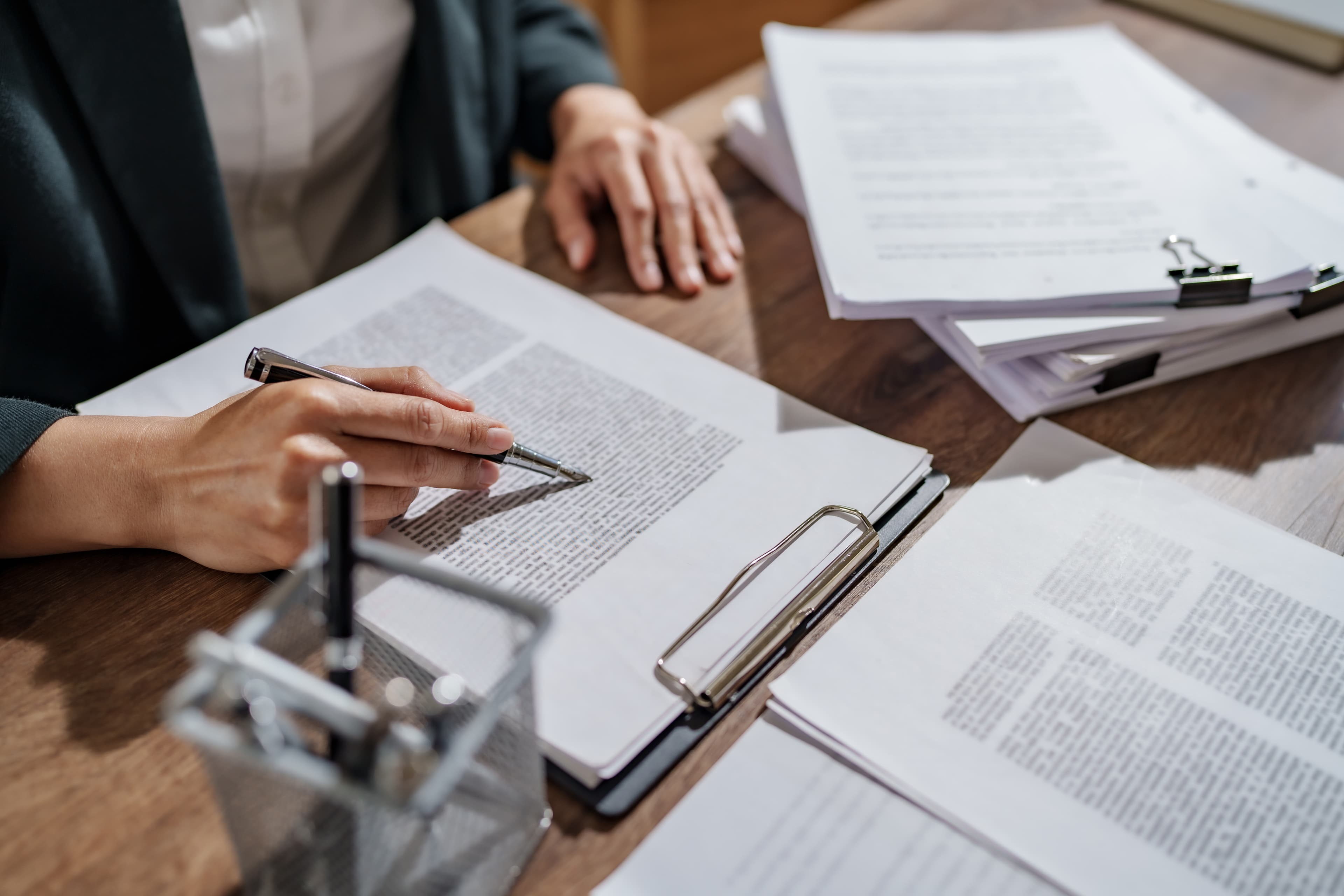 Close-up of a person reviewing printed legal documents with a pen, seated at a wooden desk with stacks of paperwork and a metal pen holder.
