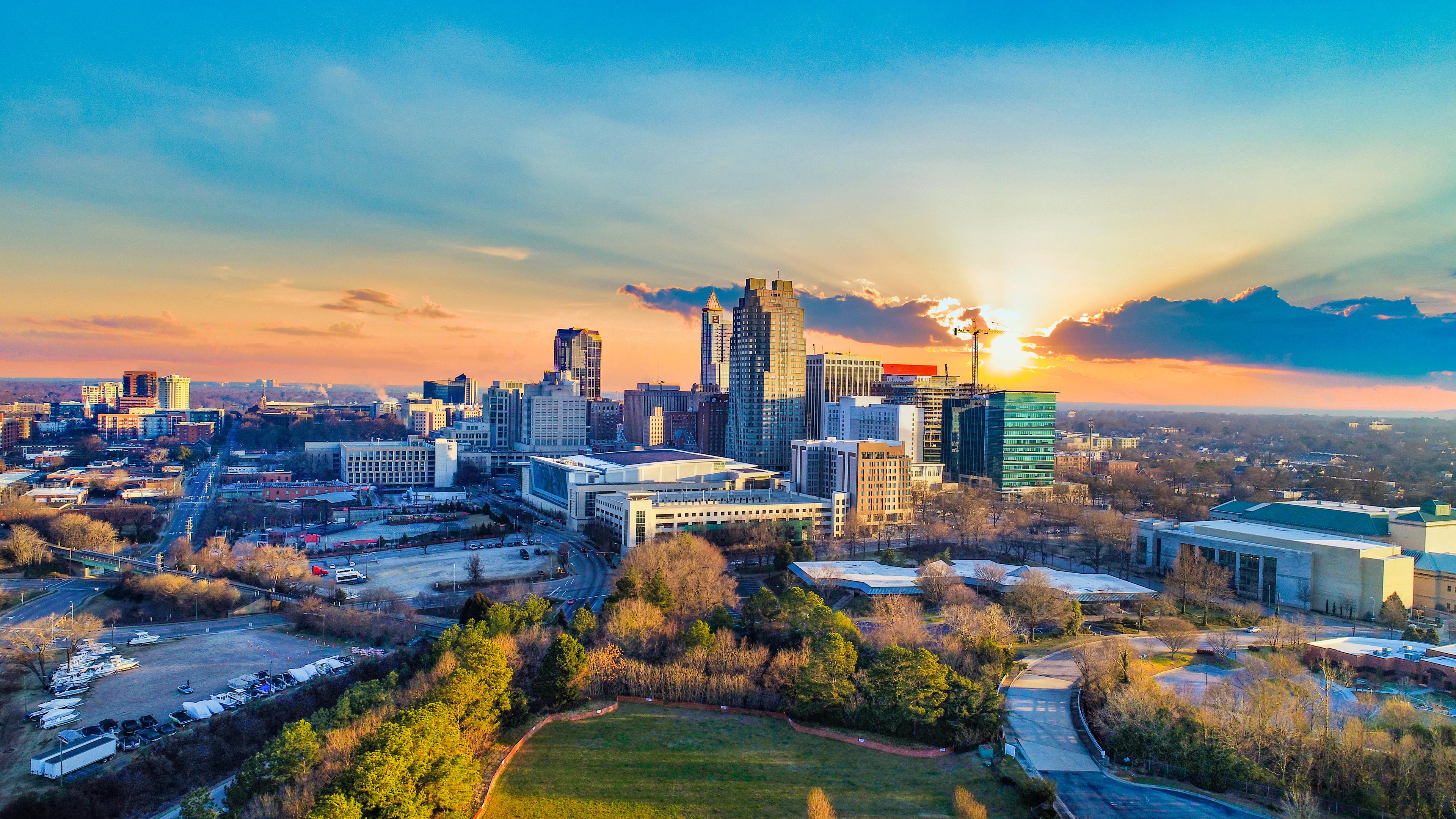 Aerial view of downtown Raleigh, North Carolina at sunset, with sunlight streaming through clouds behind the skyline and vibrant colors casting a warm glow over the cityscape.