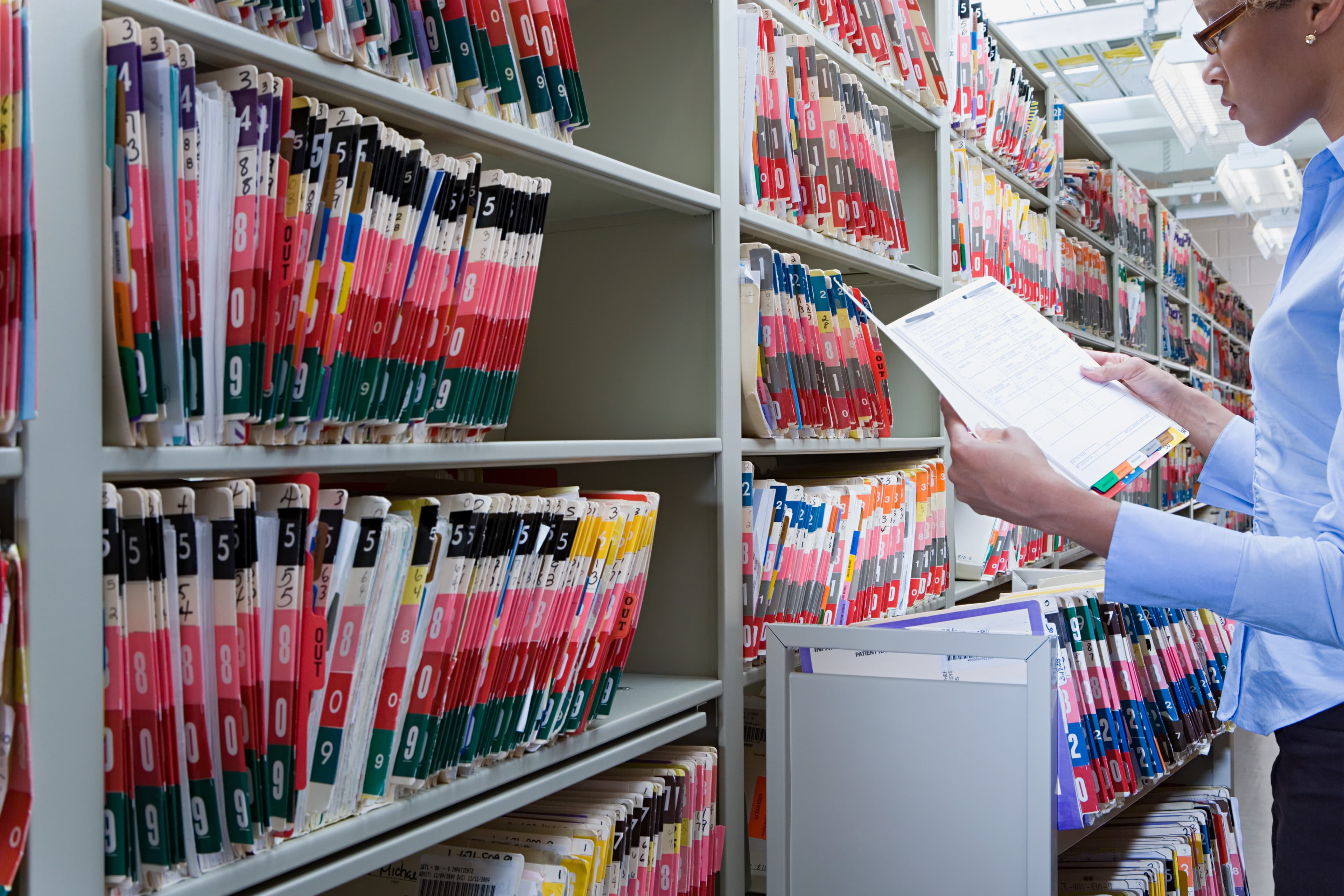 Professional reviewing a medical record in front of shelves filled with color-coded patient files in a records archive.