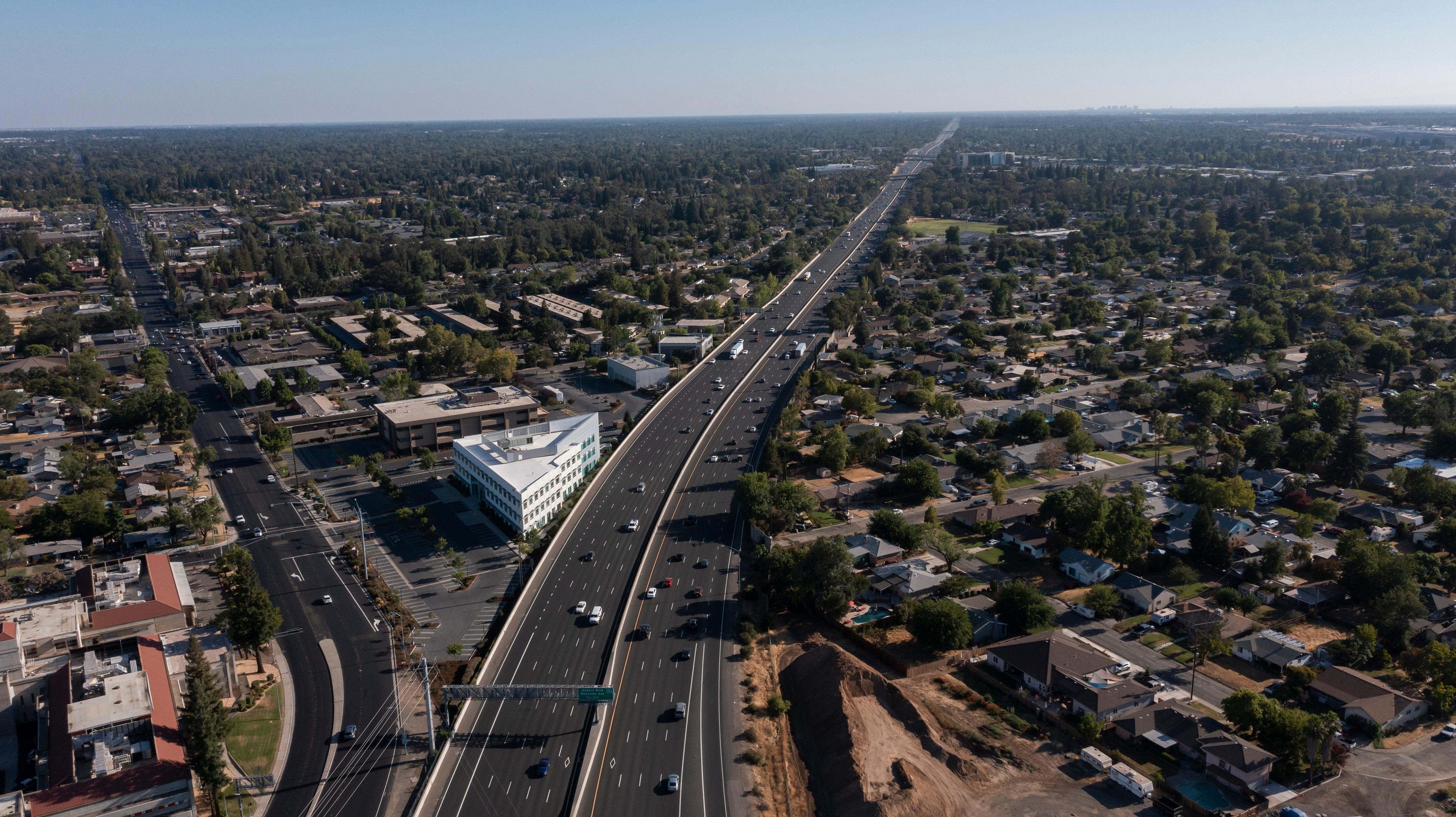 An aerial view of Roseville, California, highlighting a major highway cutting through the suburban landscape. The area is filled with residential homes, businesses, and trees, extending toward the horizon under a clear blue sky.