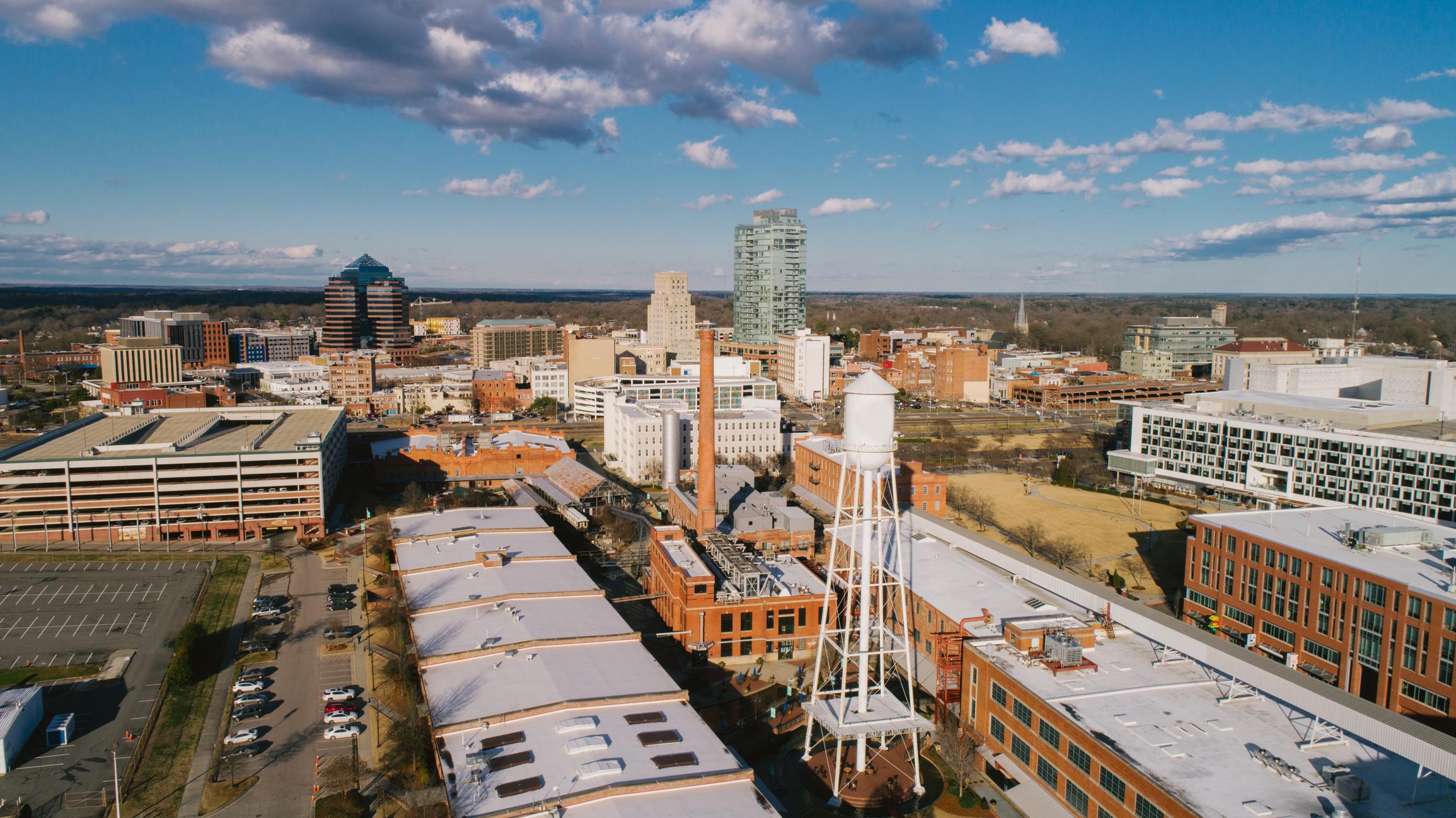 Aerial view of downtown Durham, North Carolina on a clear day, featuring the historic water tower, red-brick warehouses, and modern skyline set against a backdrop of blue sky and scattered clouds.