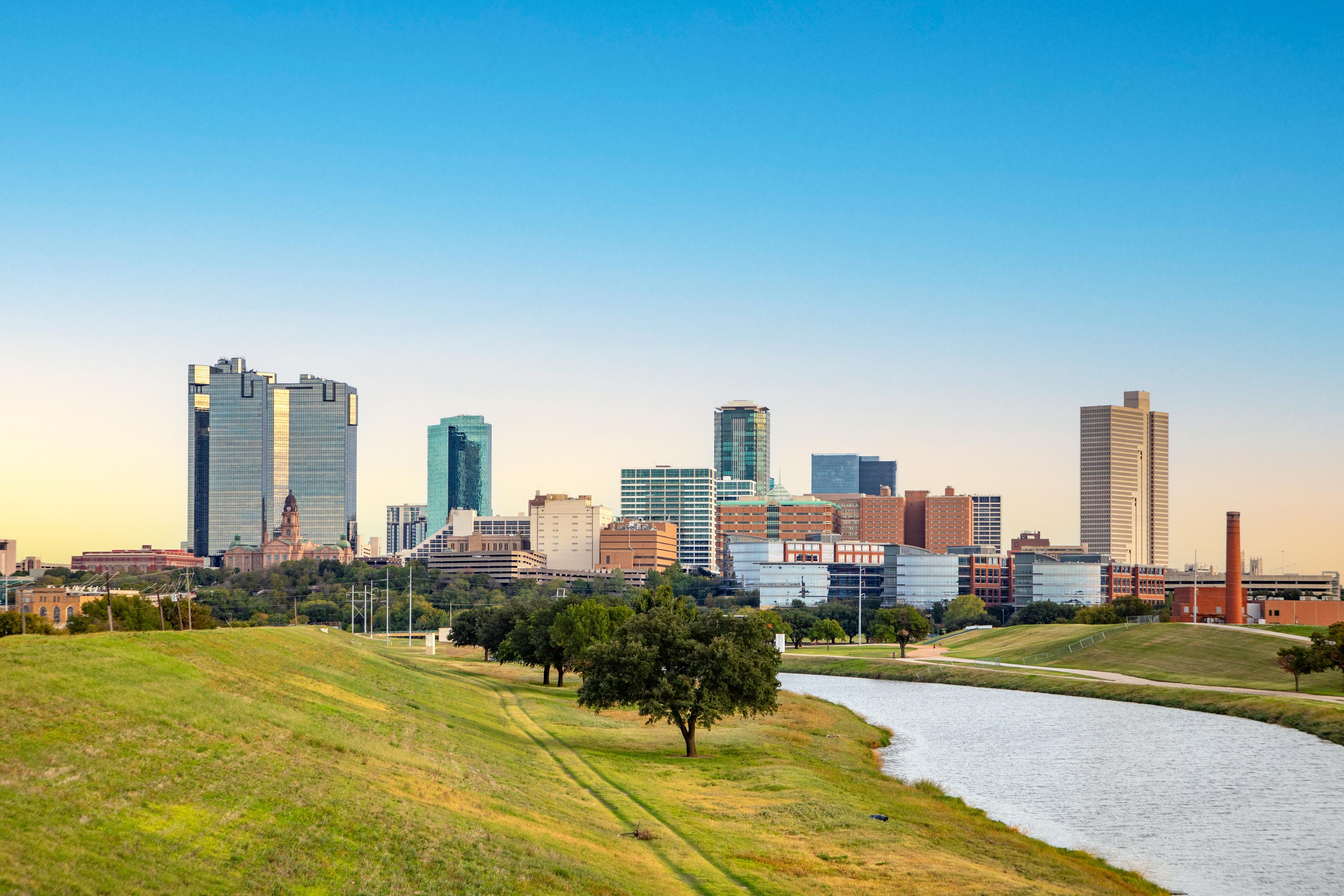 Peaceful view of the Fort Worth skyline at dusk, with modern and historic buildings rising beyond a winding river and lush green landscape in the foreground.