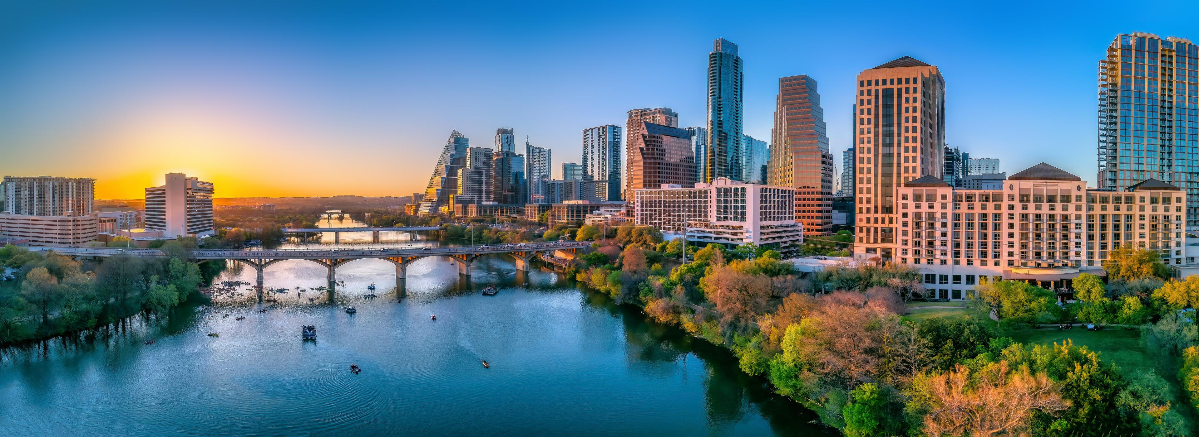 Stunning panoramic view of Austin, Texas at sunset, showcasing the city’s modern skyline, scenic riverfront, and vibrant energy as boats glide along the water.
