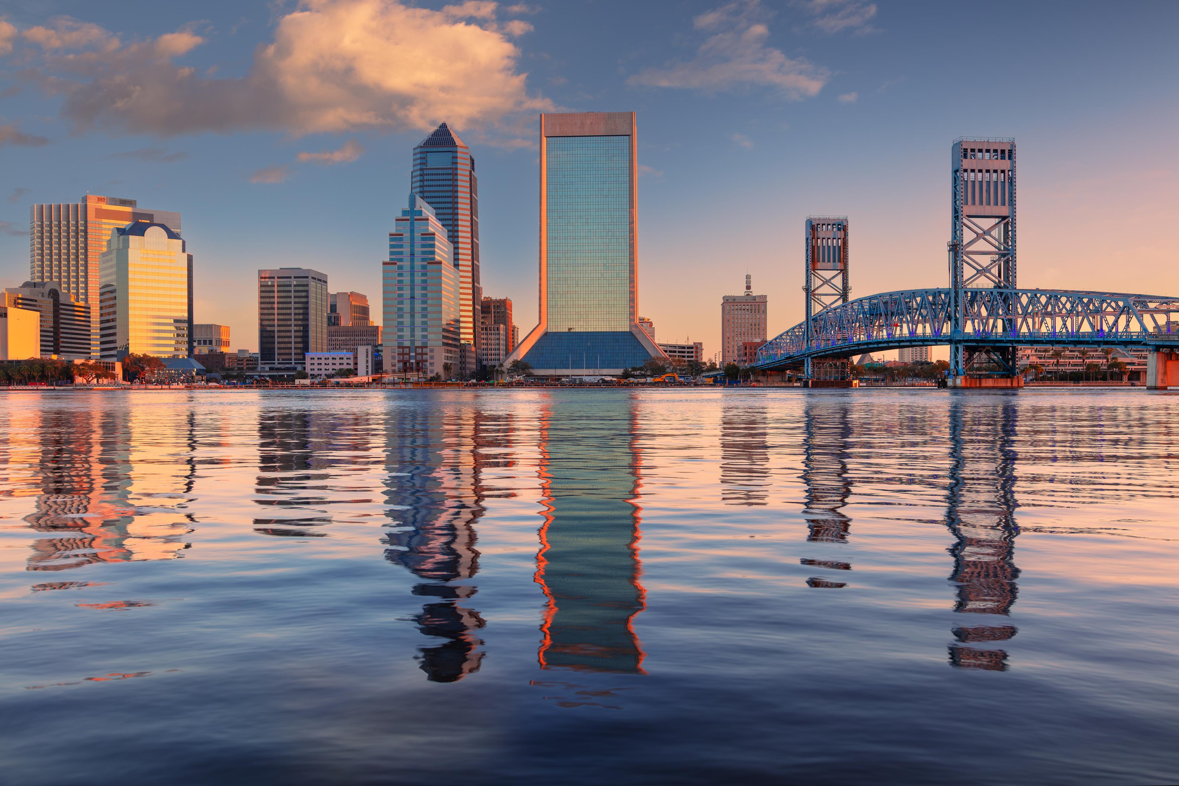 Scenic view of downtown Jacksonville, Florida, at sunset featuring the iconic Main Street Bridge and skyline reflections on the tranquil St. Johns River.