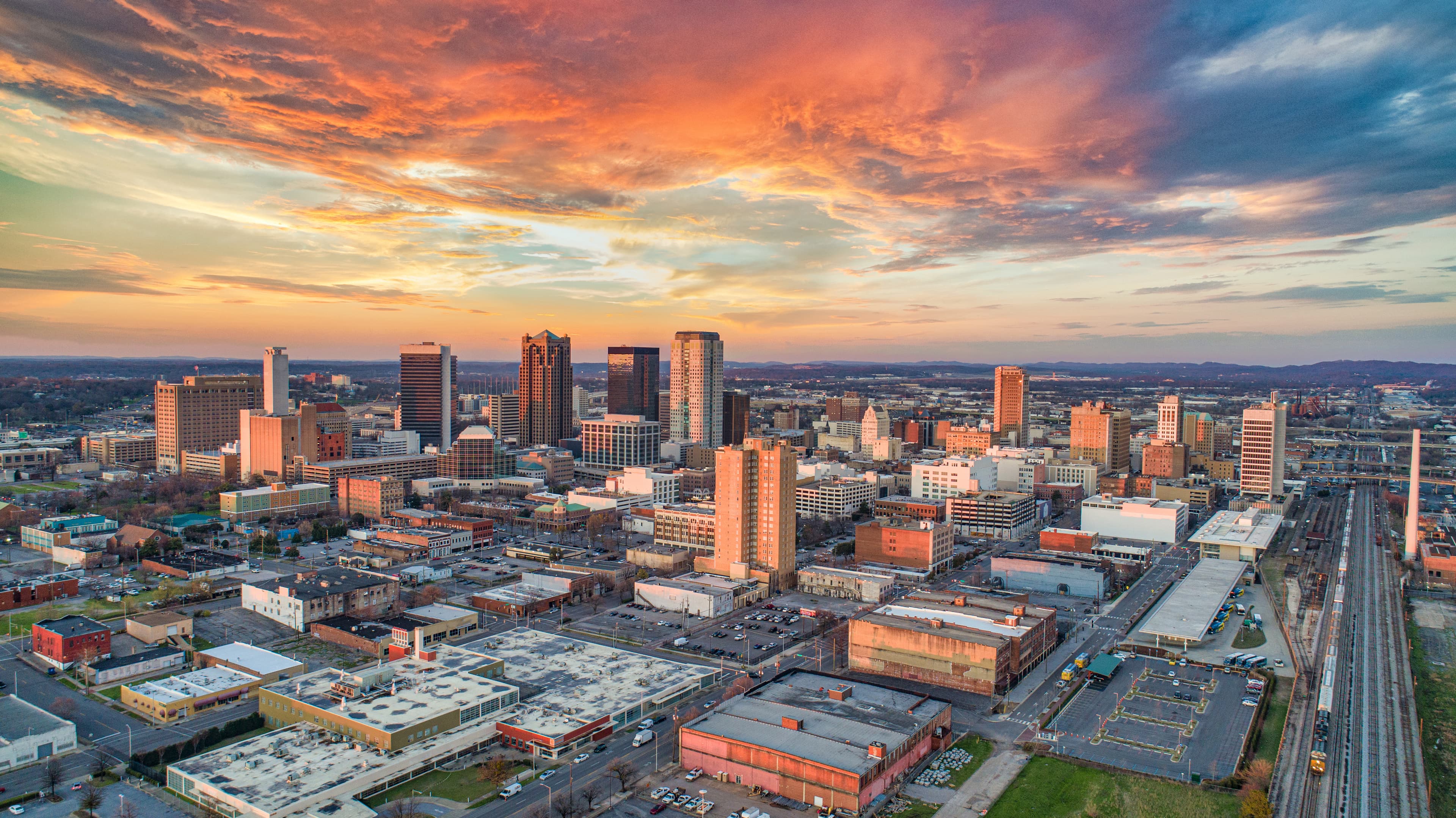 Aerial view of the downtown Birmingham, Alabama skyline at sunset with city buildings, streets and dramatic orange clouds.