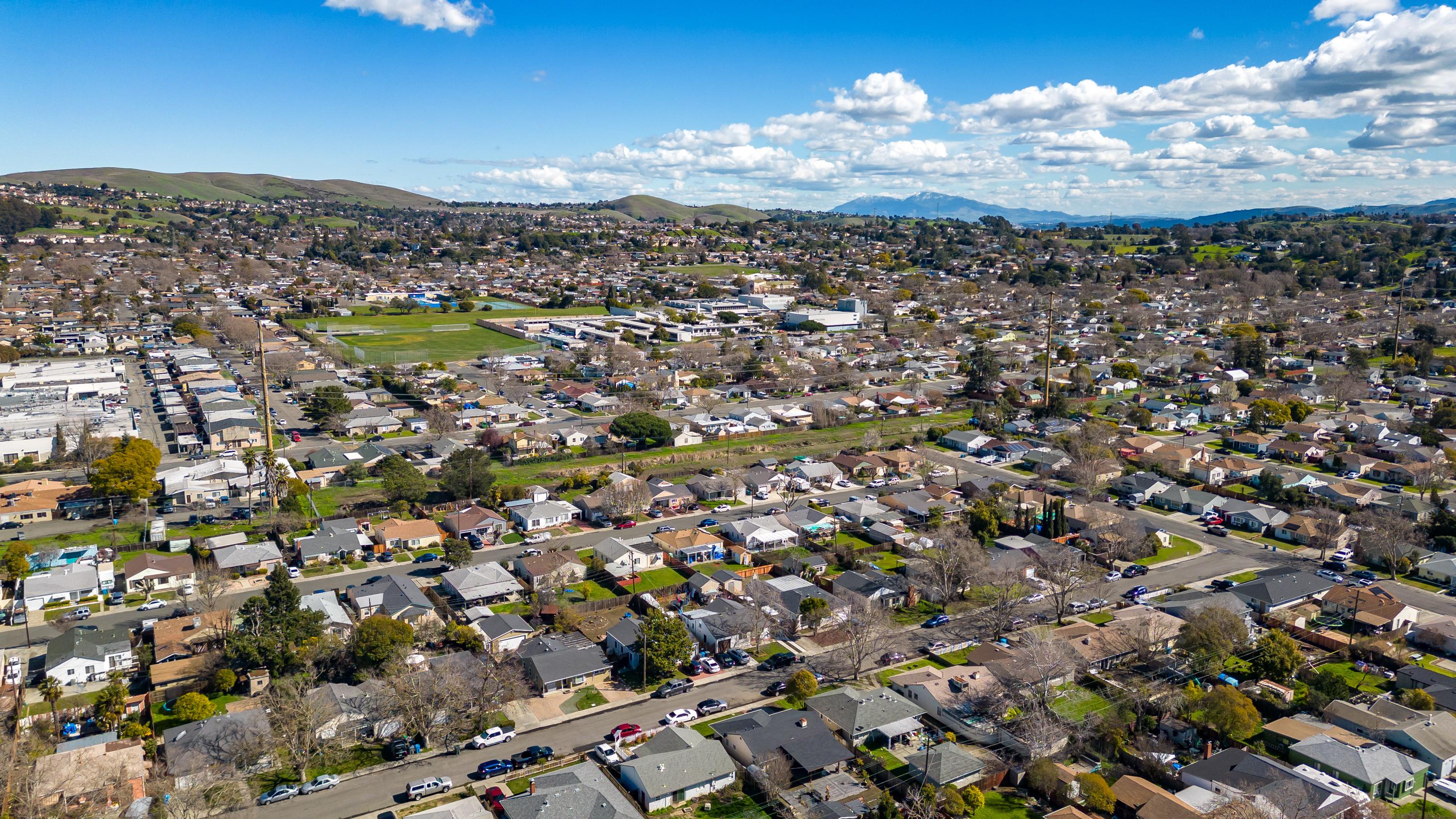 An aerial view of Vallejo, California, showcasing a residential neighborhood with neatly arranged houses, tree-lined streets, and surrounding green hills. A school campus and open fields are visible in the mid-ground, while distant rolling hills and a clear blue sky with scattered clouds add to the scenic backdrop.