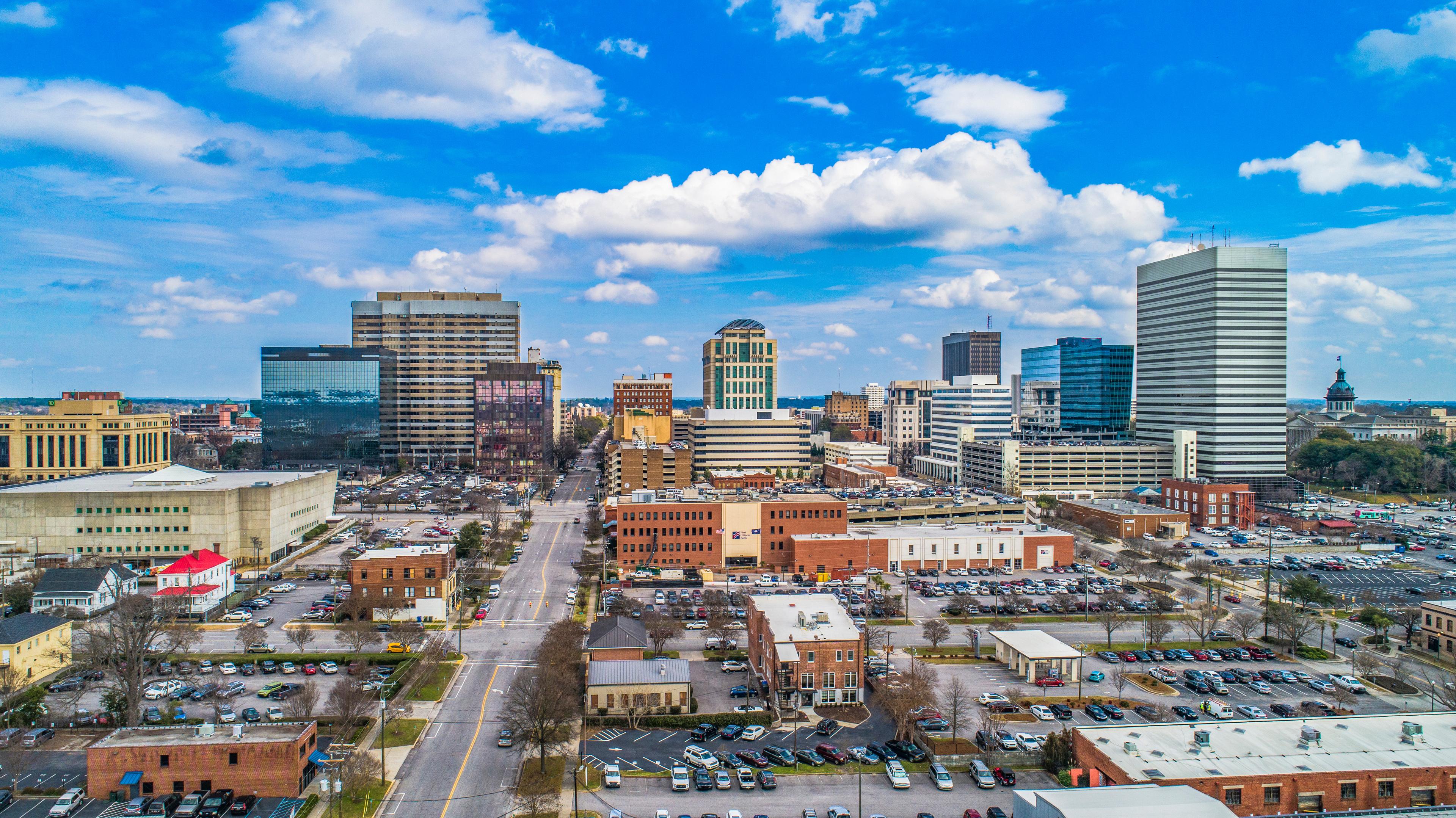 Aerial view of downtown Columbia, South Carolina on a bright day, with a mix of modern and historic buildings under a vibrant blue sky with scattered clouds.