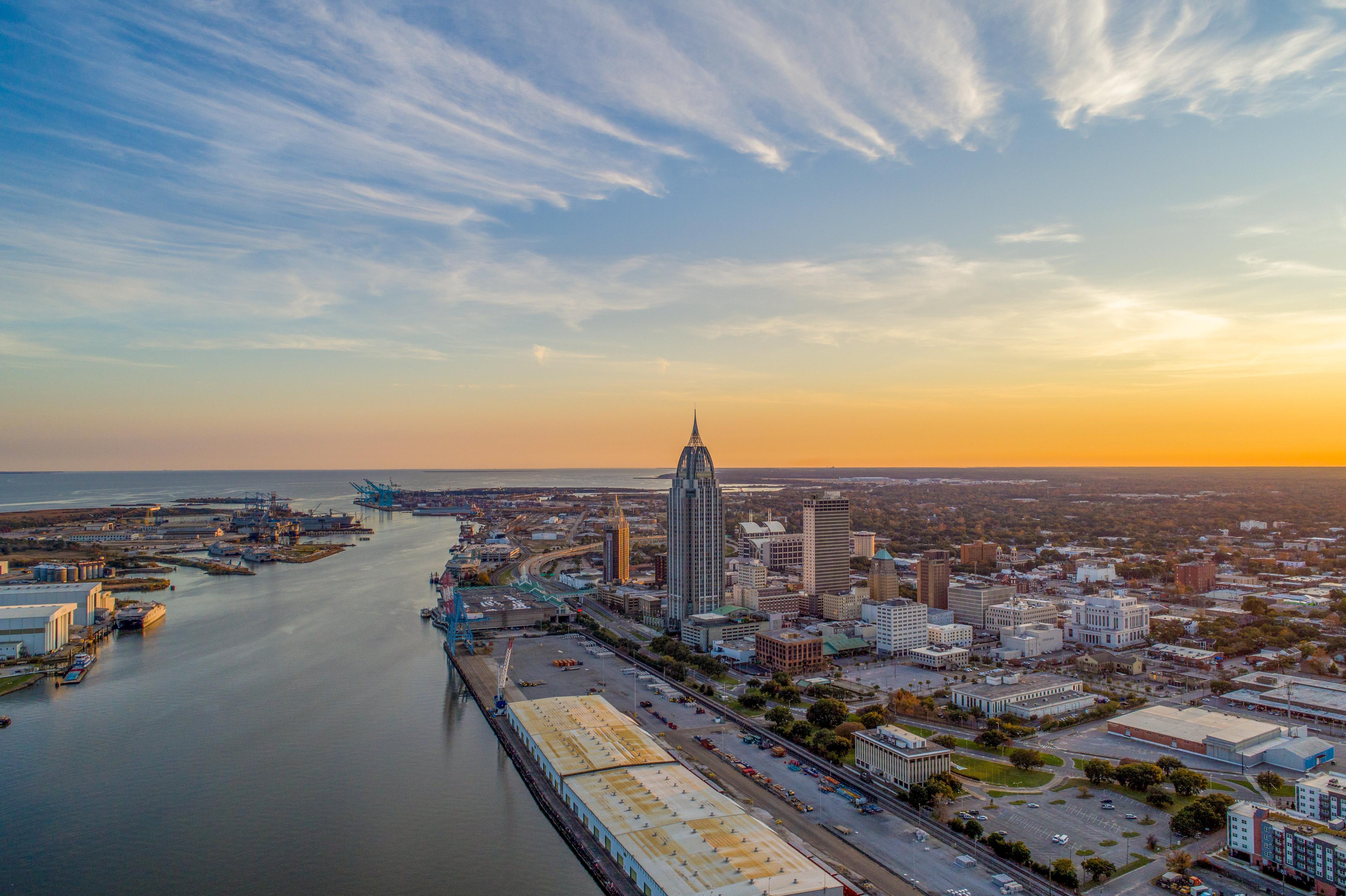 Aerial sunset view of downtown Mobile, Alabama, showcasing the skyline along the Mobile River with the RSA Battle House Tower and busy port facilities under a glowing Gulf Coast sky.