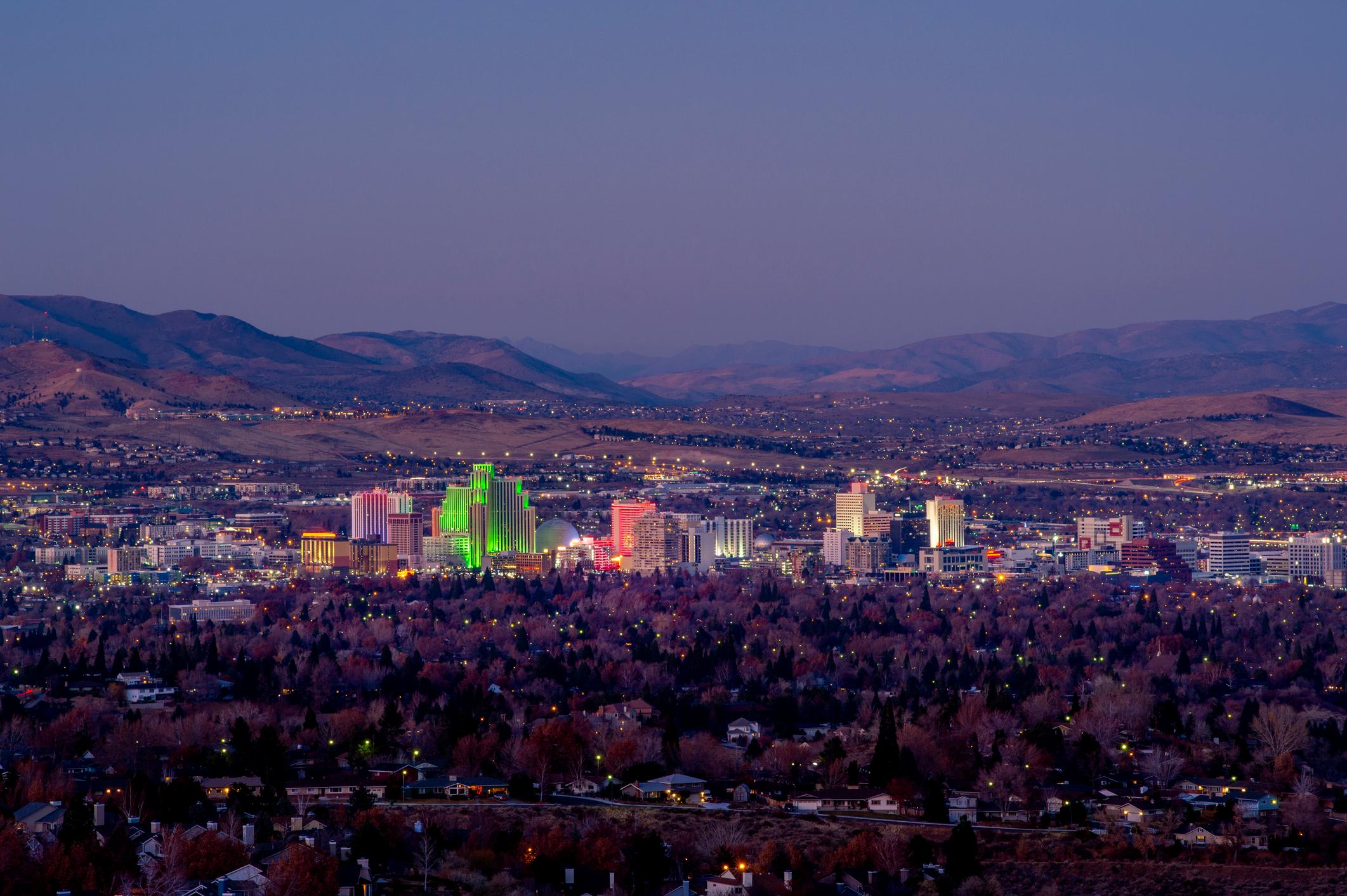 A twilight aerial view of Reno, Nevada, showcasing a glowing cityscape with brightly lit buildings in various colors. The scene is framed by dark trees in the foreground and rolling desert hills under a fading evening sky.