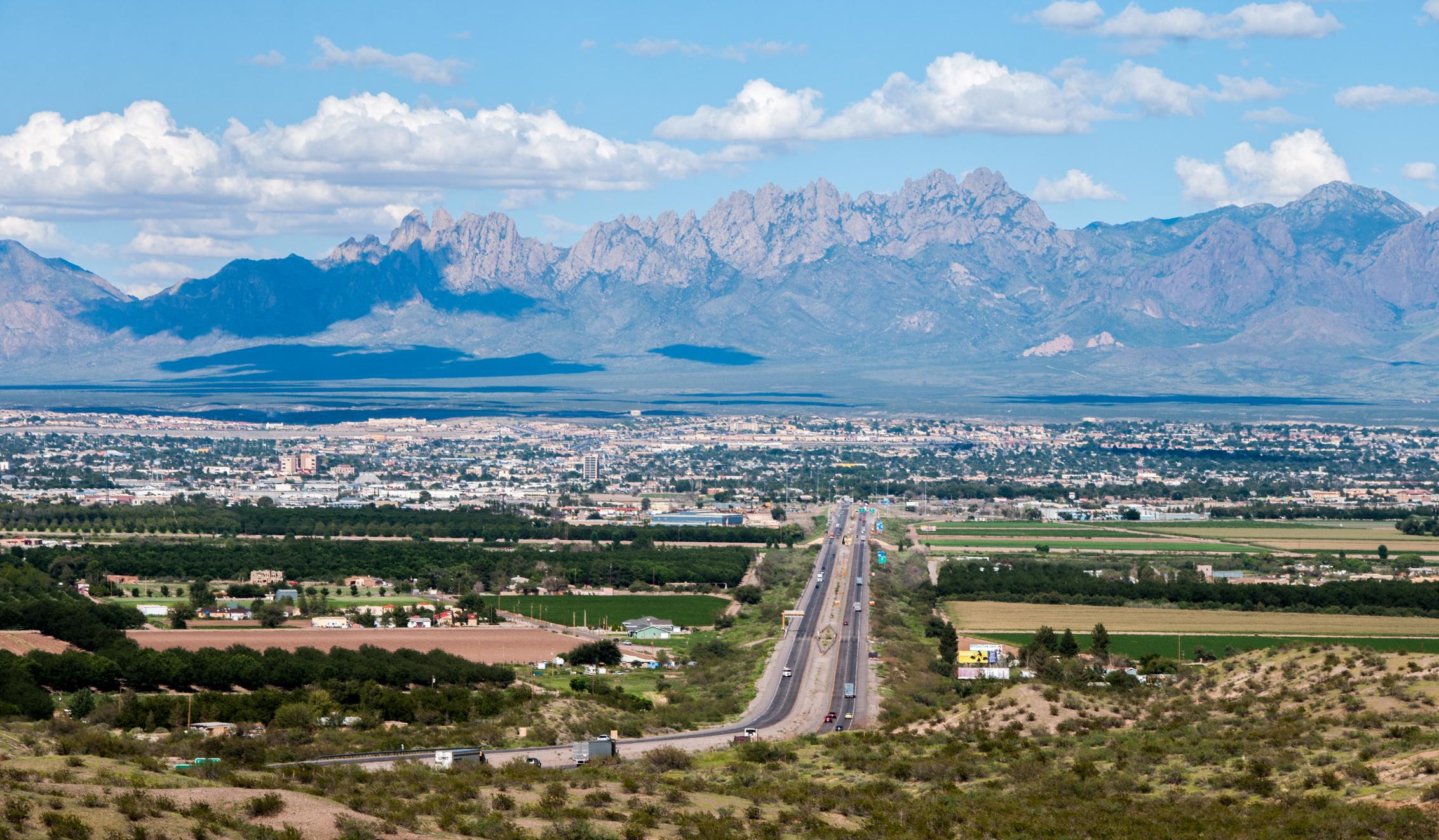 A scenic view of Las Cruces, New Mexico, with the Organ Mountains in the background, a bustling highway leading into the city, and vibrant farmland in the foreground under a bright blue sky with scattered clouds.