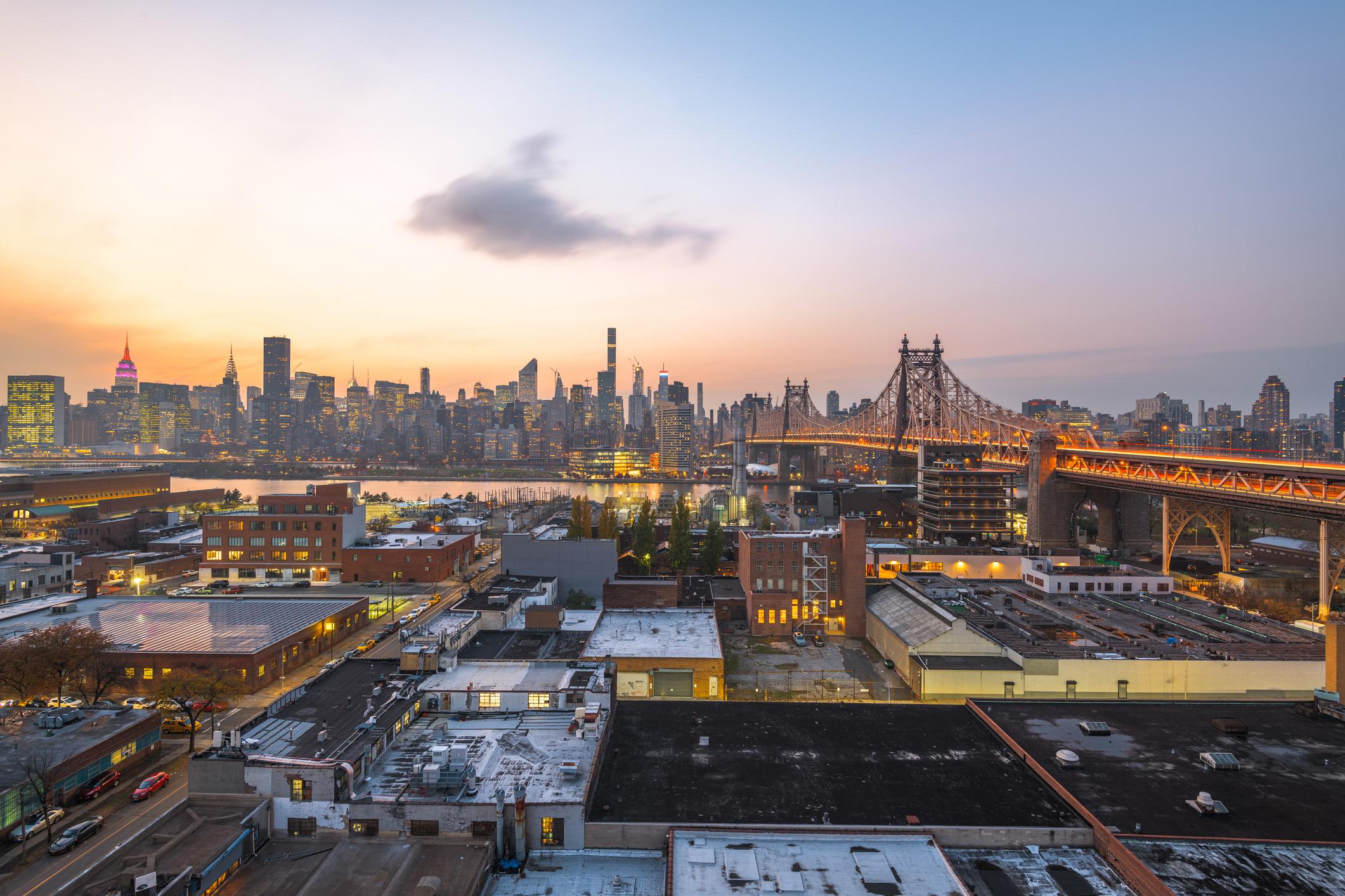 A view of Queens, New York, at sunset, featuring the Queensboro Bridge illuminated with warm lights stretching across the East River. In the foreground, industrial rooftops and red-brick buildings are visible, while the Manhattan skyline, with the Empire State Building lit in pink and other iconic skyscrapers, creates a backdrop under a pastel sky.