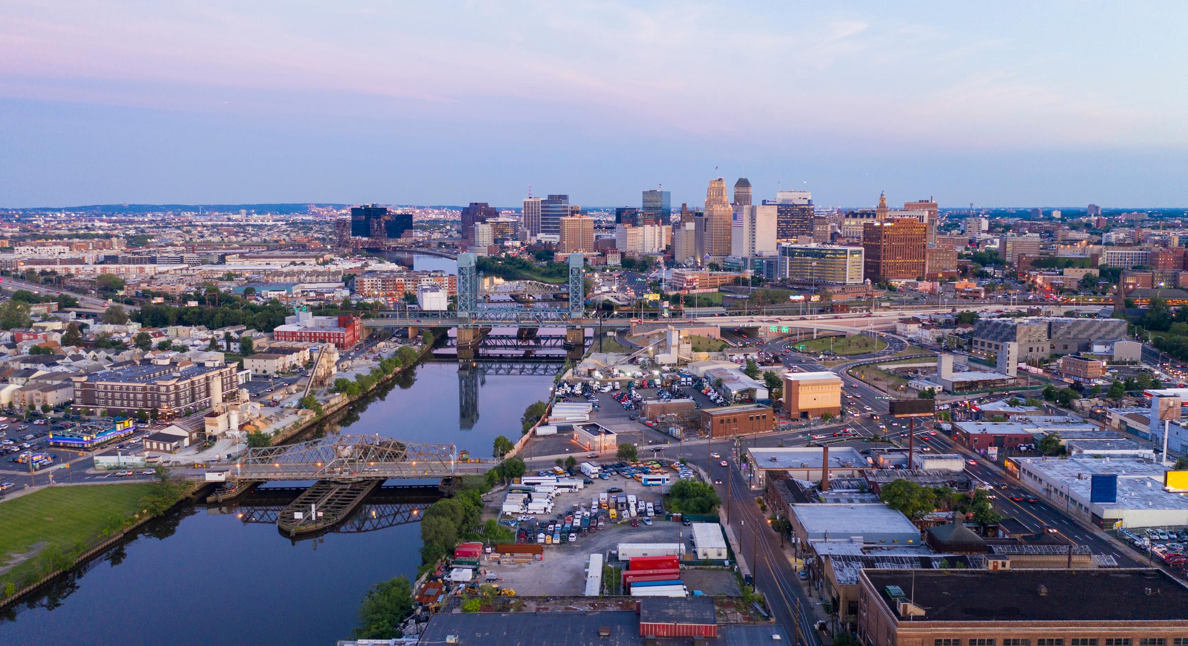 A wide-angle aerial view of Newark, New Jersey, showing the city skyline with a mix of modern and historic buildings. The Passaic River cuts through the scene, lined by bridges, residential areas, and industrial zones, with highways and busy streets stretching into the distance under a soft, colorful sky.