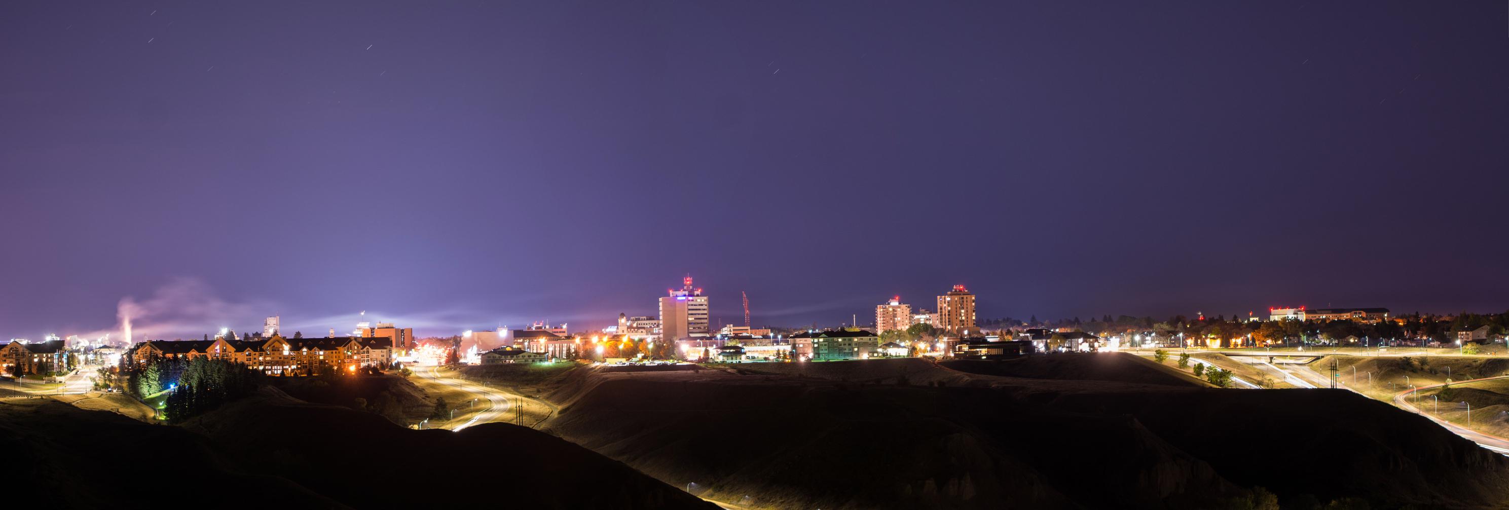 A panoramic night view of Lethbridge, Alberta, featuring illuminated buildings and winding roads, with a clear dark sky enhancing the city's lights.