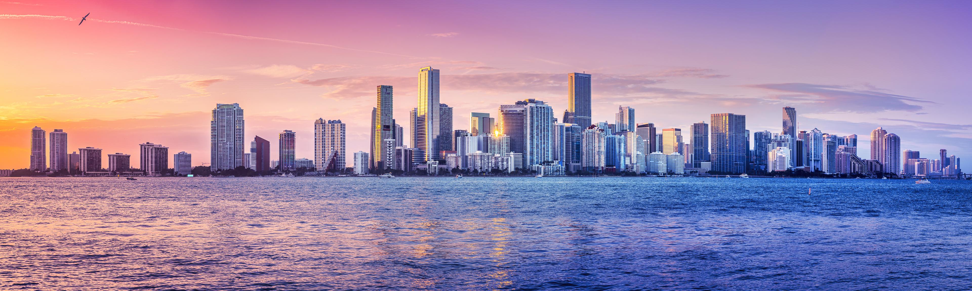 Panoramic view of Miami's modern skyline at sunset, with high-rise buildings reflecting the warm glow over the calm waters of Biscayne Bay.