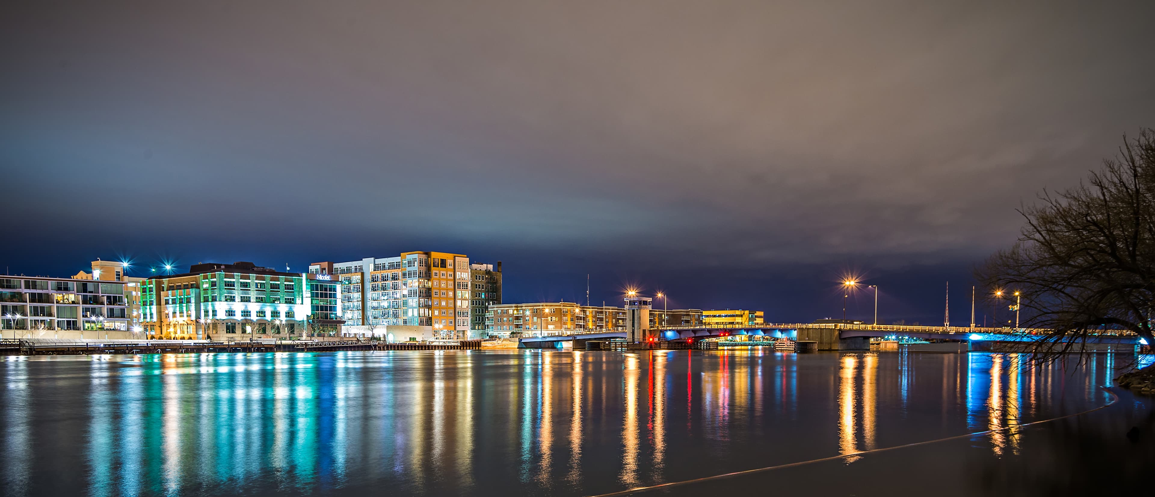 Downtown Green Bay, Wisconsin waterfront at night with illuminated buildings, a bridge and city lights reflecting across the Fox River.