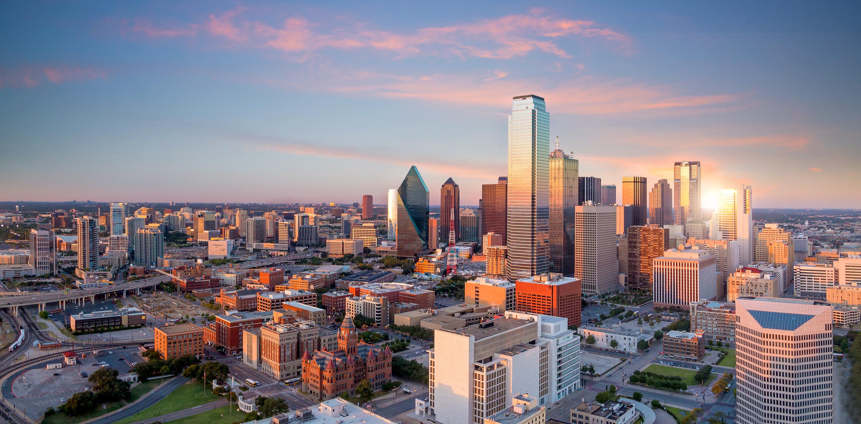 Aerial view of downtown Dallas at sunset, showcasing the vibrant cityscape and modern architecture under a colorful sky.