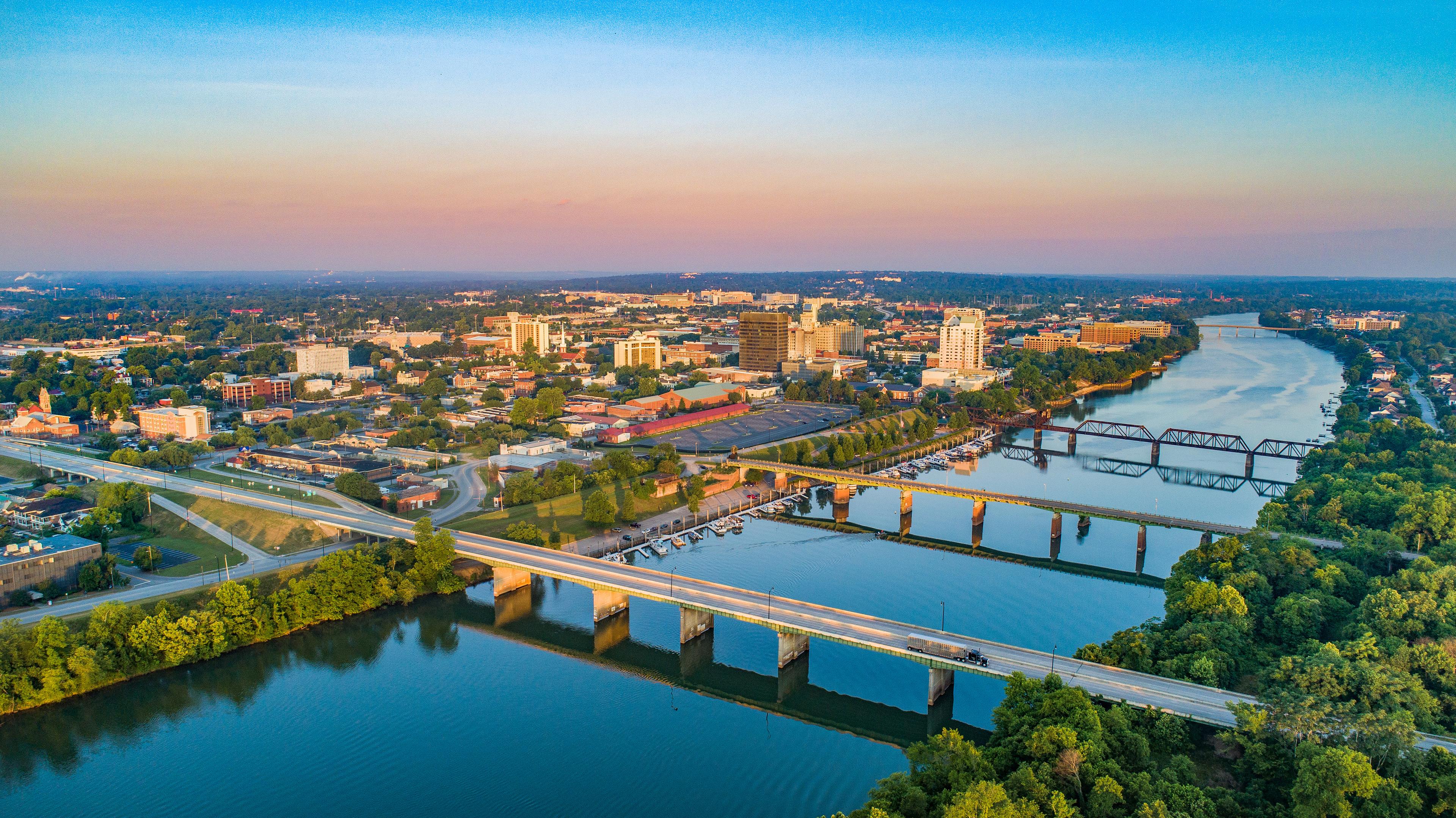 Scenic aerial view of downtown Augusta, Georgia at sunset, with bridges spanning the Savannah River and the city skyline framed by golden light and lush greenery.
