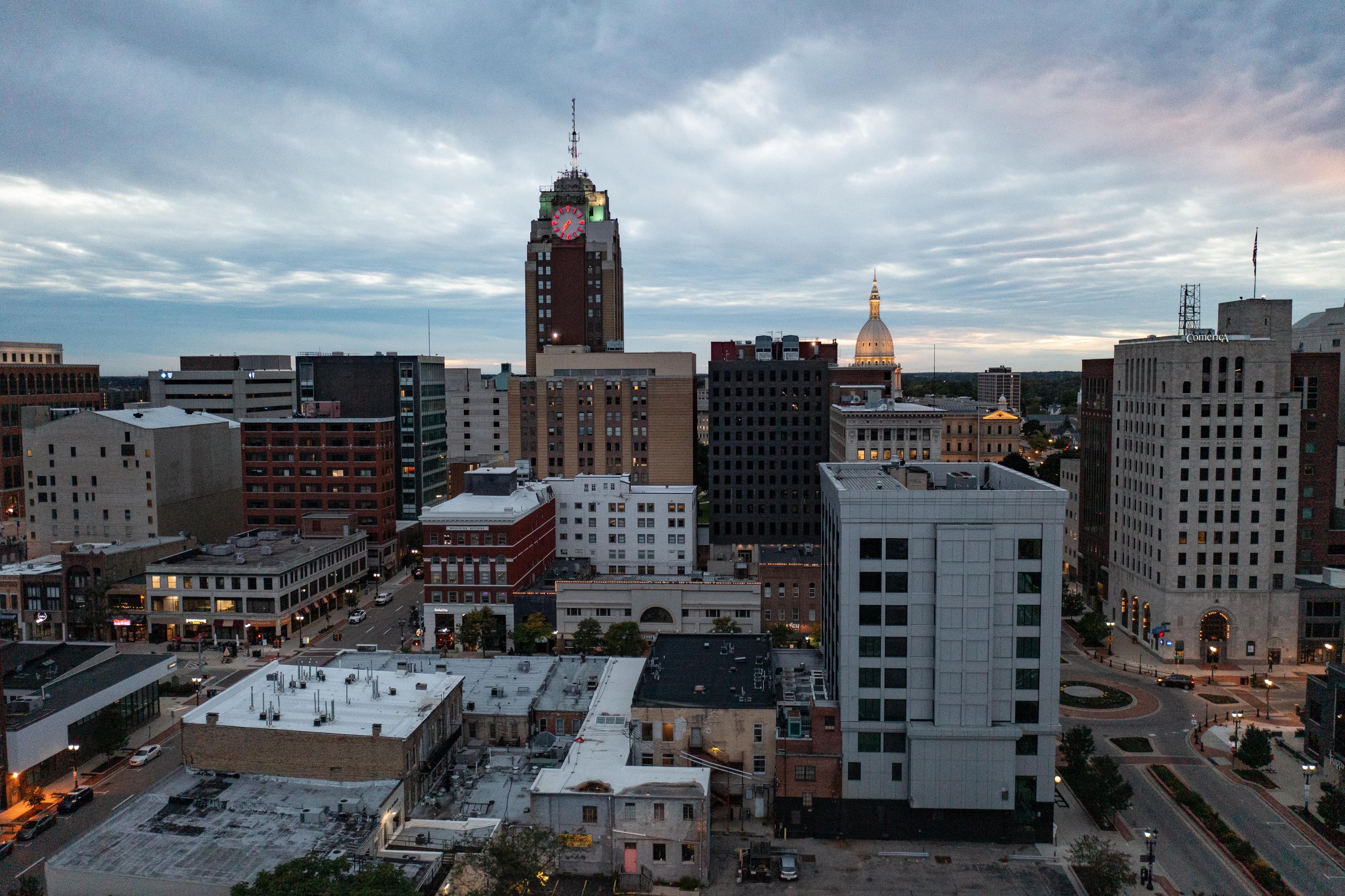 Downtown Lansing, Michigan at dusk, featuring mid-rise office buildings, the iconic Boji Tower with a glowing red clock, and the Michigan State Capitol dome illuminated in the distance under a cloudy sky.