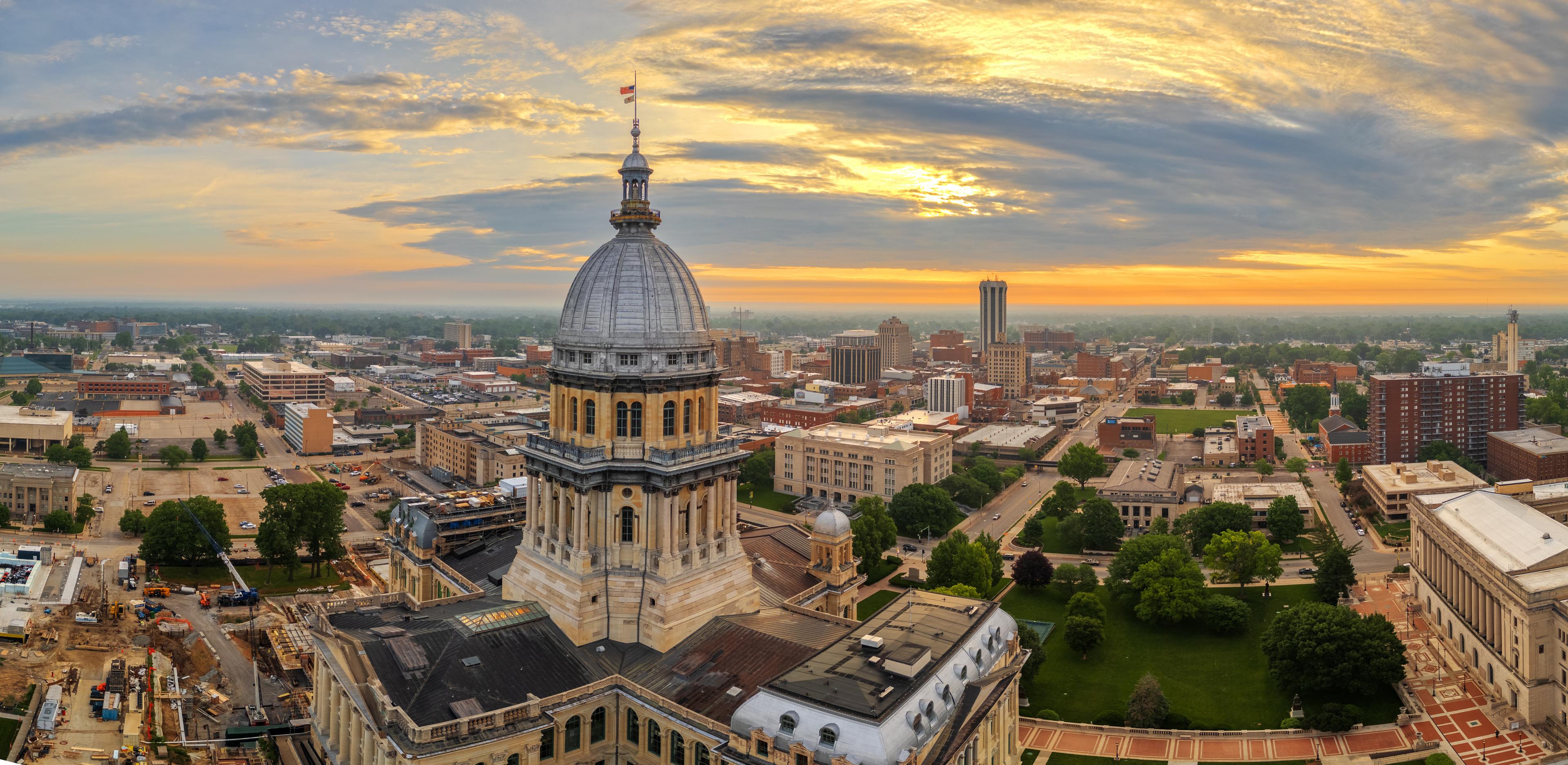 Drone view of the Illinois State Capitol at sunrise, with downtown Springfield’s skyline stretching into the distance under a glowing, cloud-filled sky.
