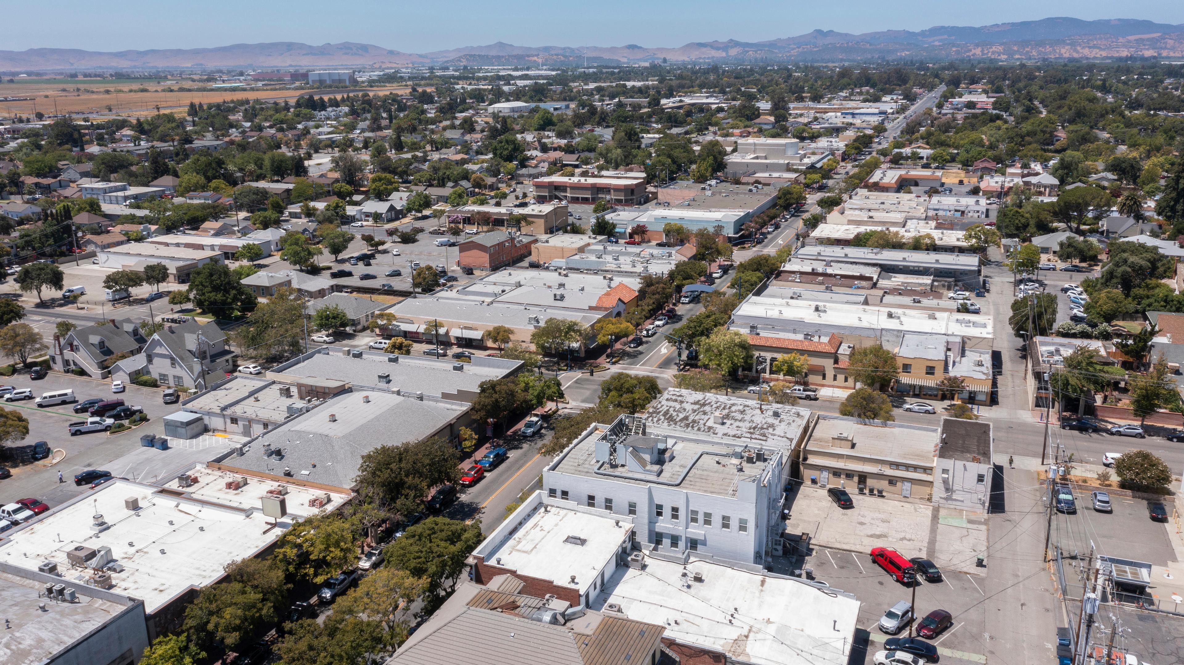 An aerial view of downtown Fairfield, California, showcasing a mix of residential and commercial buildings, tree-lined streets, and distant hills under a bright blue sky.