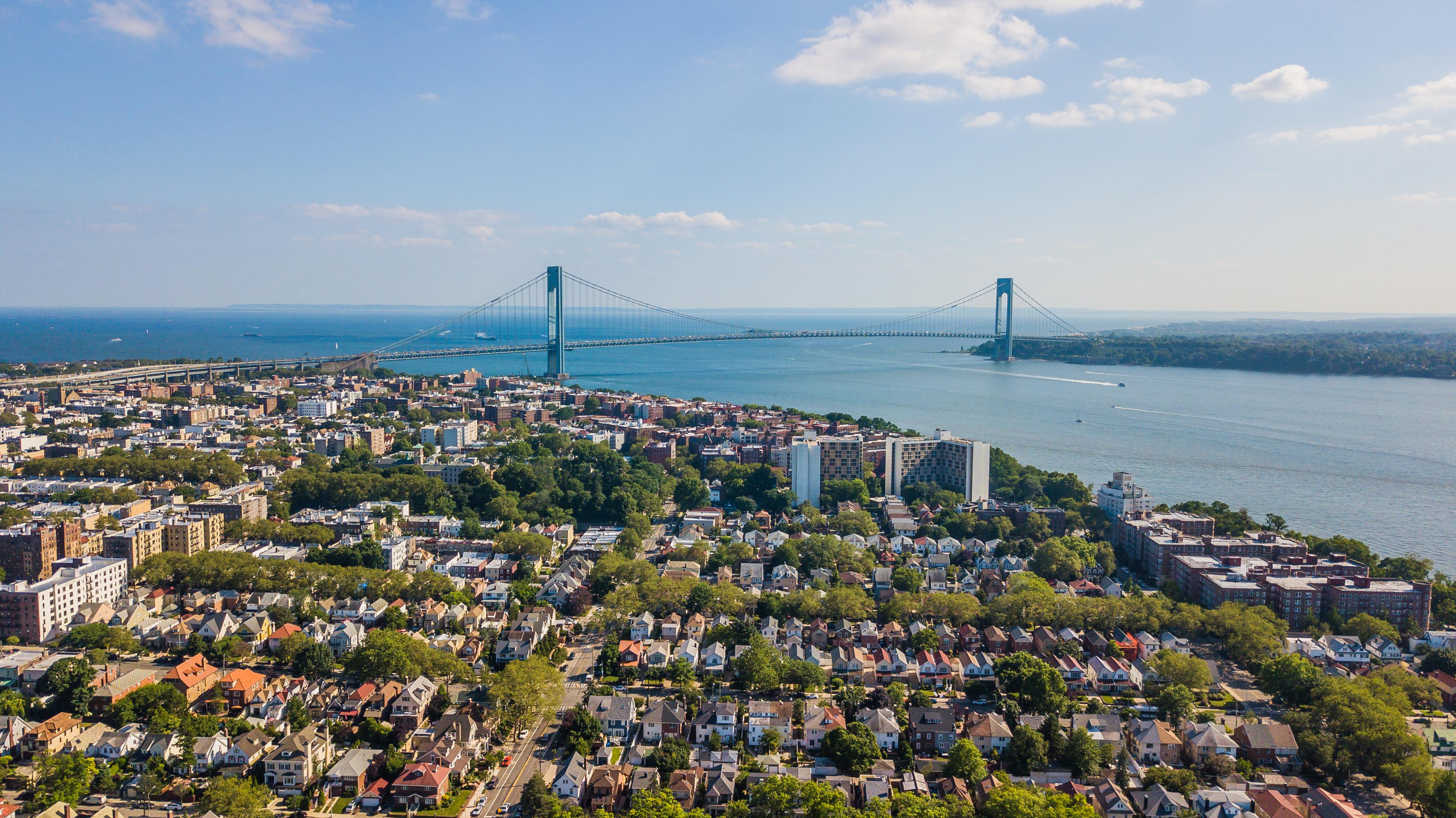 An aerial view of Brooklyn, New York, featuring a neighborhood with rows of houses and apartment buildings surrounded by green trees. In the distance, the Verrazzano-Narrows Bridge stretches across the water under a clear blue sky, connecting Brooklyn to Staten Island.