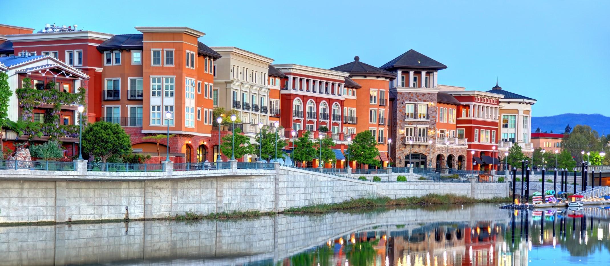 A vibrant waterfront view in Napa, California, featuring colorful Mediterranean-style buildings reflected on the calm water, with street lamps and lush greenery lining the promenade.