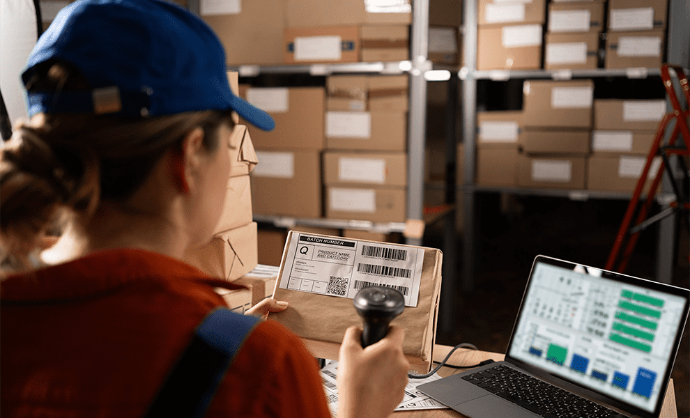 A woman in a warehouse standing near a desk with a data dashboard displayed on a laptop, scanning a barcode on a box. 


