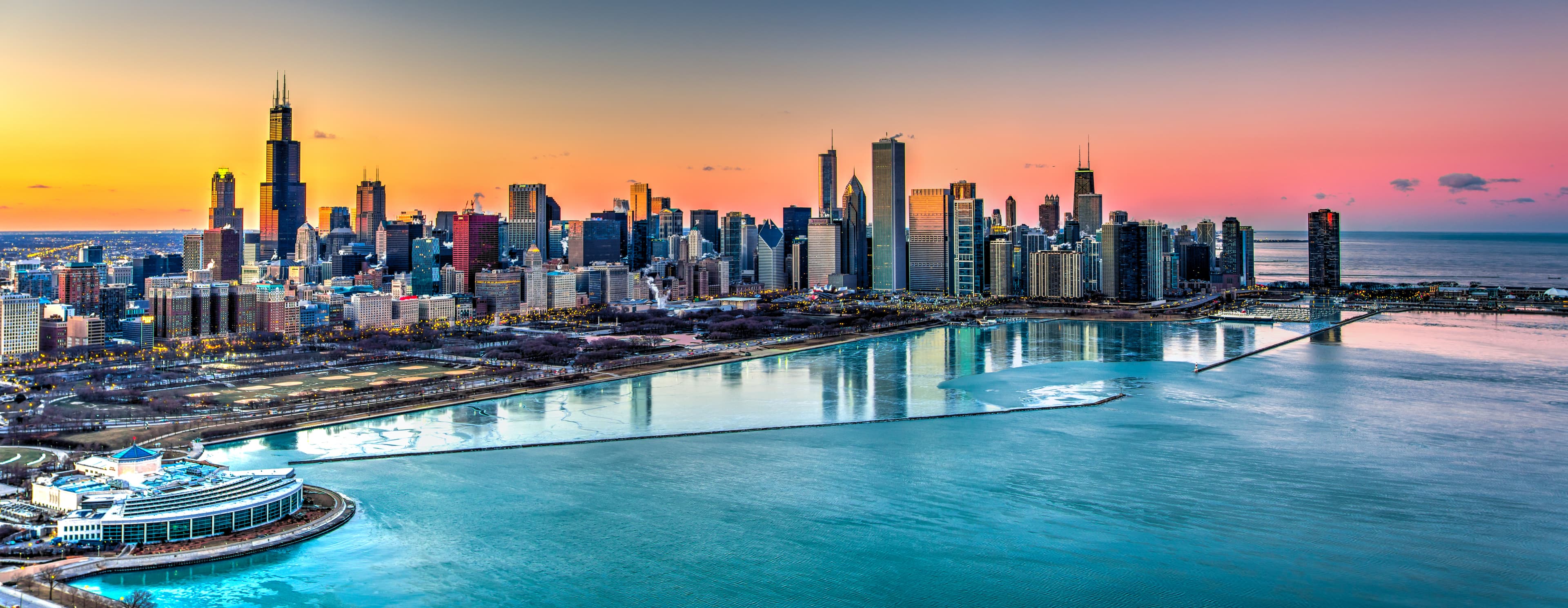 Aerial view of the Chicago skyline at sunset, featuring the Willis Tower and other iconic skyscrapers reflecting in Lake Michigan’s icy waters.