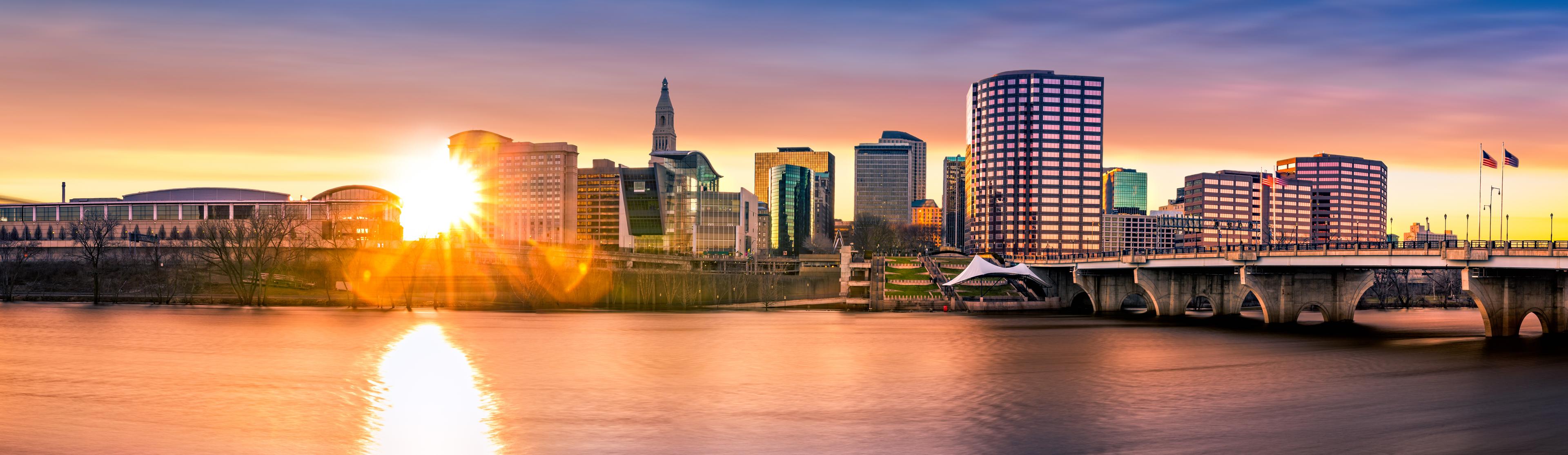 A sunset view of Hartford, Connecticut, with the skyline illuminated by golden light, the Connecticut River reflecting the warm hues, and a bridge spanning the scene.