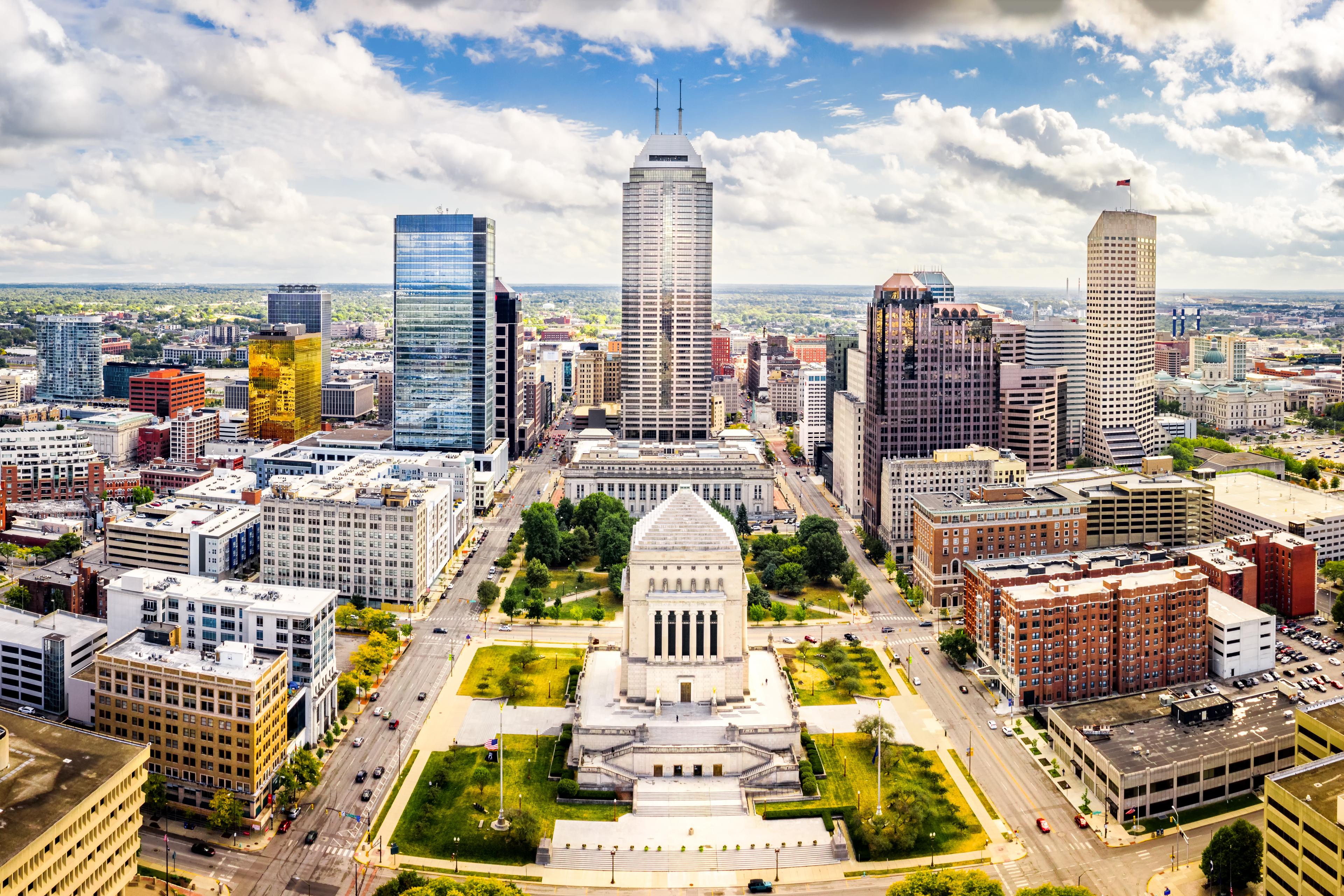 Aerial view of downtown Indianapolis showcasing the Indiana War Memorial and surrounding modern and historic skyscrapers on a bright, partly cloudy day.