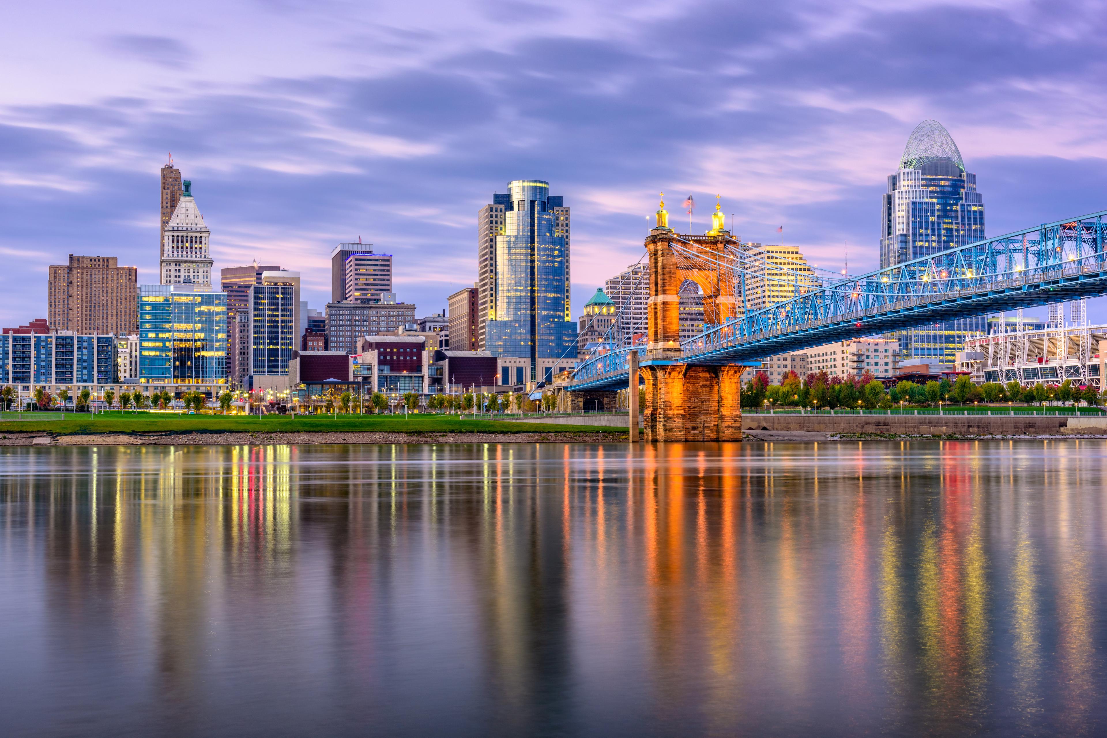 Twilight view of downtown Cincinnati, Ohio, with the illuminated John A. Roebling Suspension Bridge and city skyline reflecting in the calm waters of the Ohio River.