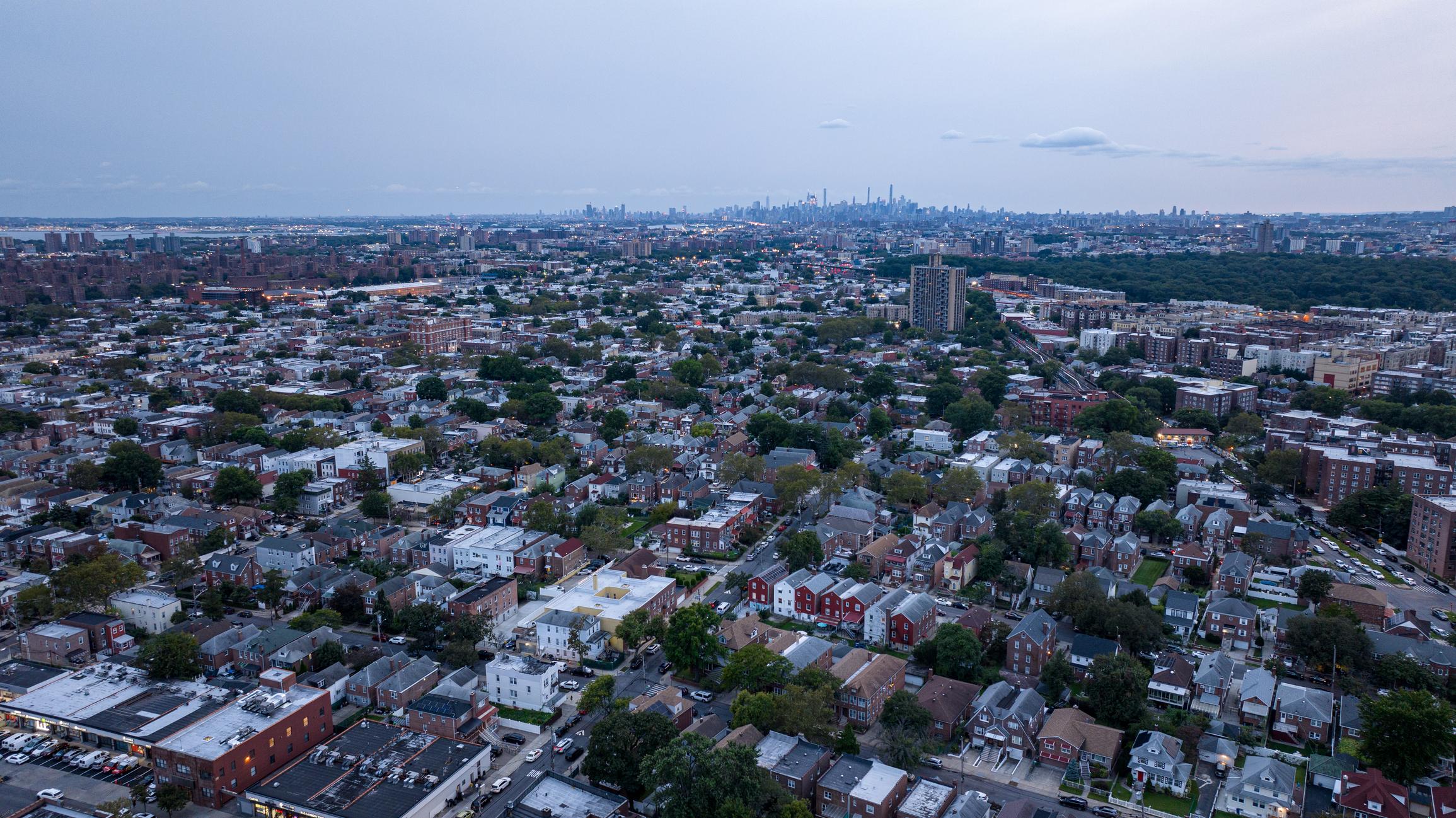 A wide aerial view of the Bronx, New York, featuring a mix of houses, apartment complexes, and tree-lined streets. The distant Manhattan skyline stretches across the horizon under a cloudy sky.