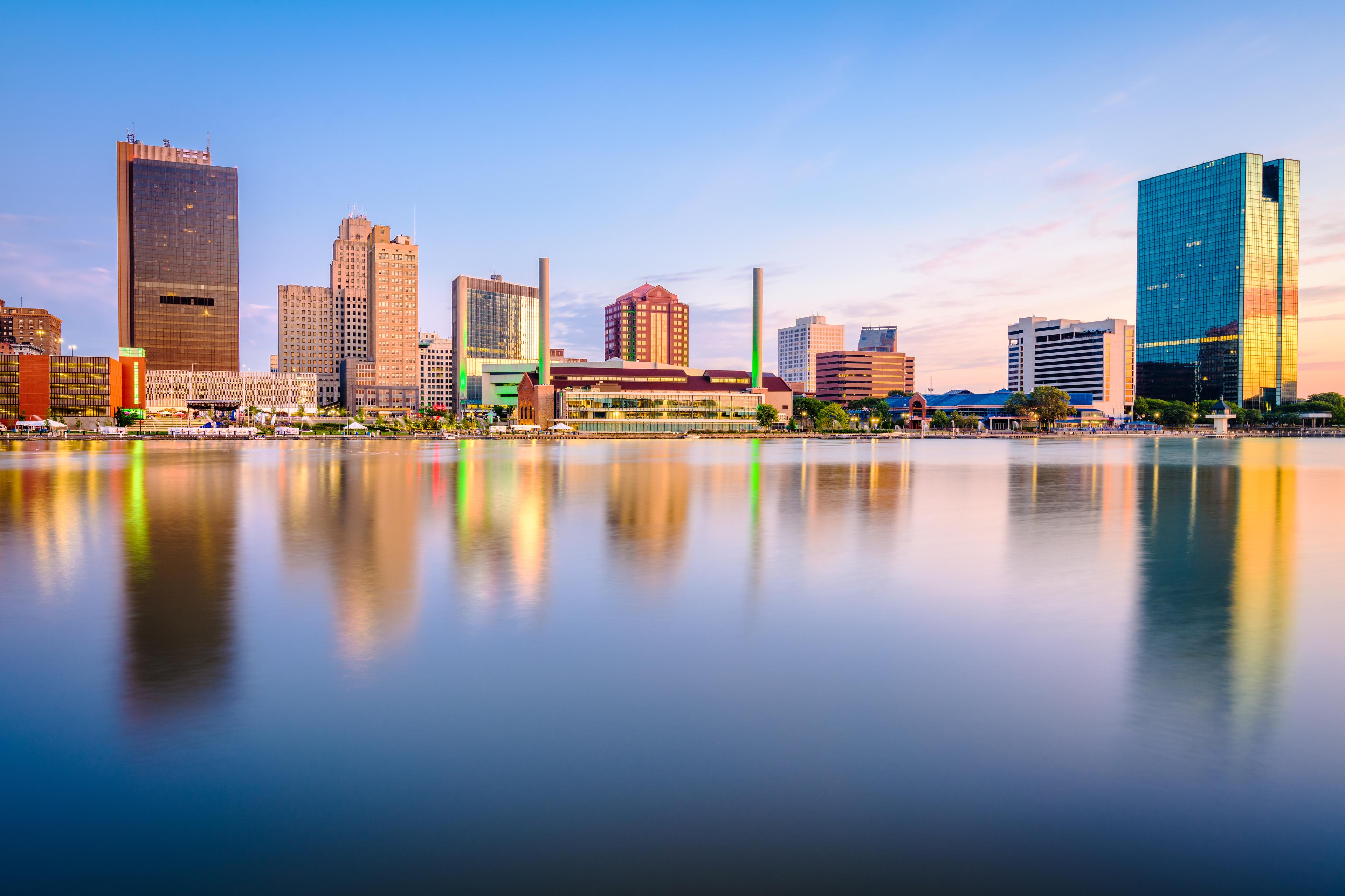 Downtown Toledo skyline reflected in the Maumee River during sunset, featuring modern office buildings and vibrant city lights.