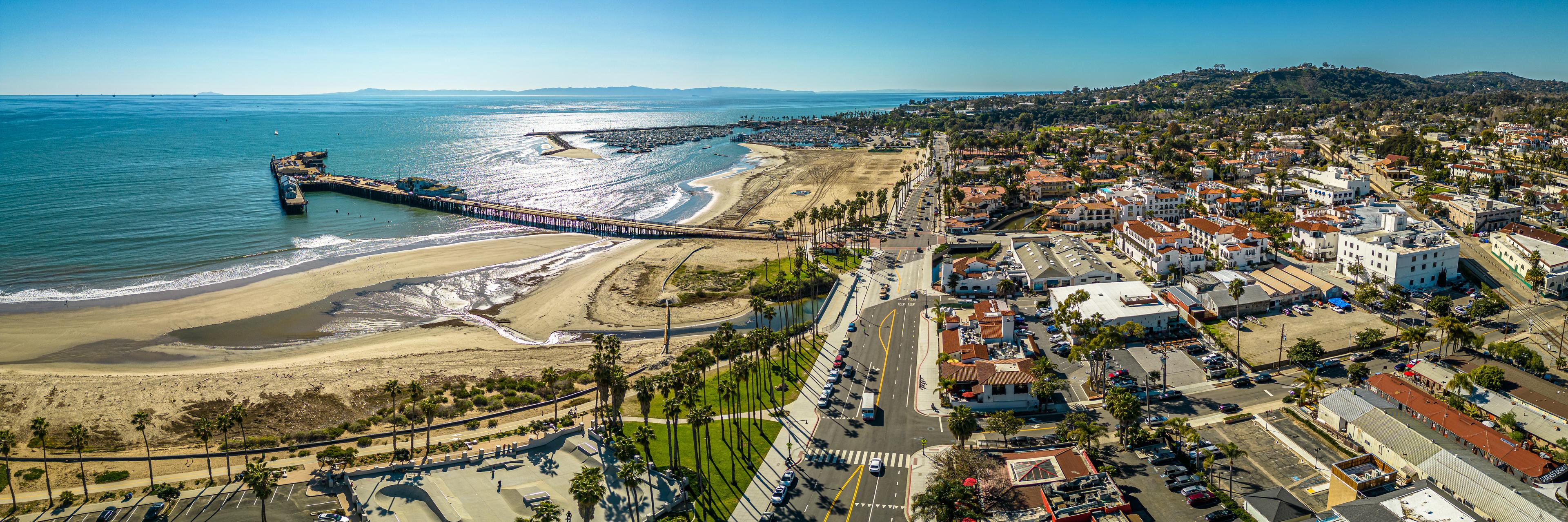 Panoramic aerial view of Santa Barbara, California, showcasing Stearns Wharf, the sandy coastline, and red-tiled roofs of the city against a backdrop of sparkling ocean and coastal hills.