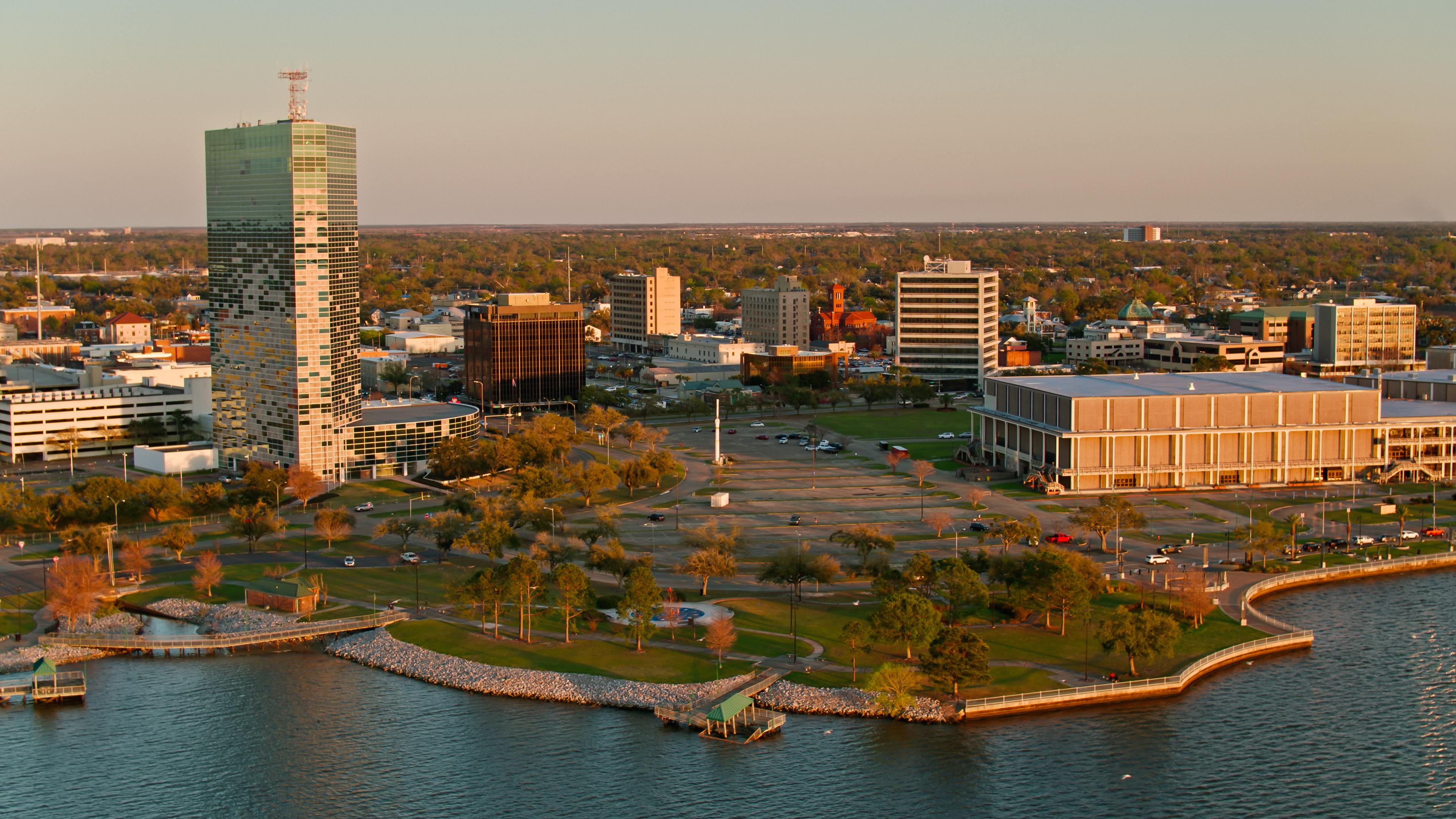 An aerial view of downtown Lake Charles, Louisiana, showcasing the iconic Capital One Tower, the Civic Center, and the waterfront park area under warm evening light.