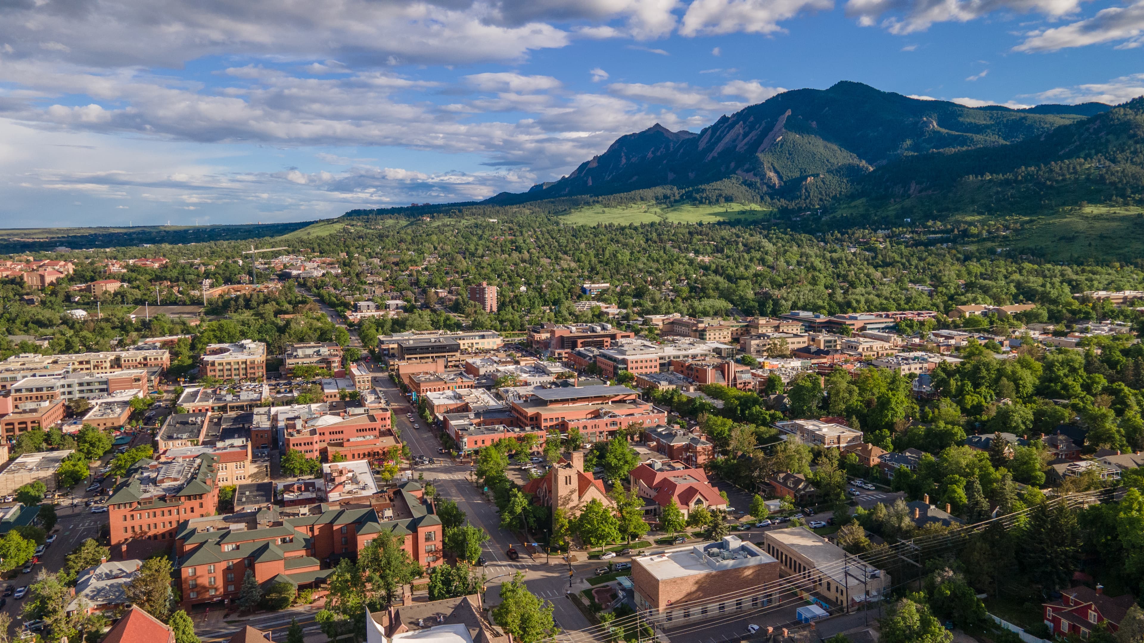Aerial view of Boulder, Colorado, showing a blend of commercial and residential buildings surrounded by dense greenery, with the iconic Flatirons mountain range rising in the background under a partly cloudy blue sky.