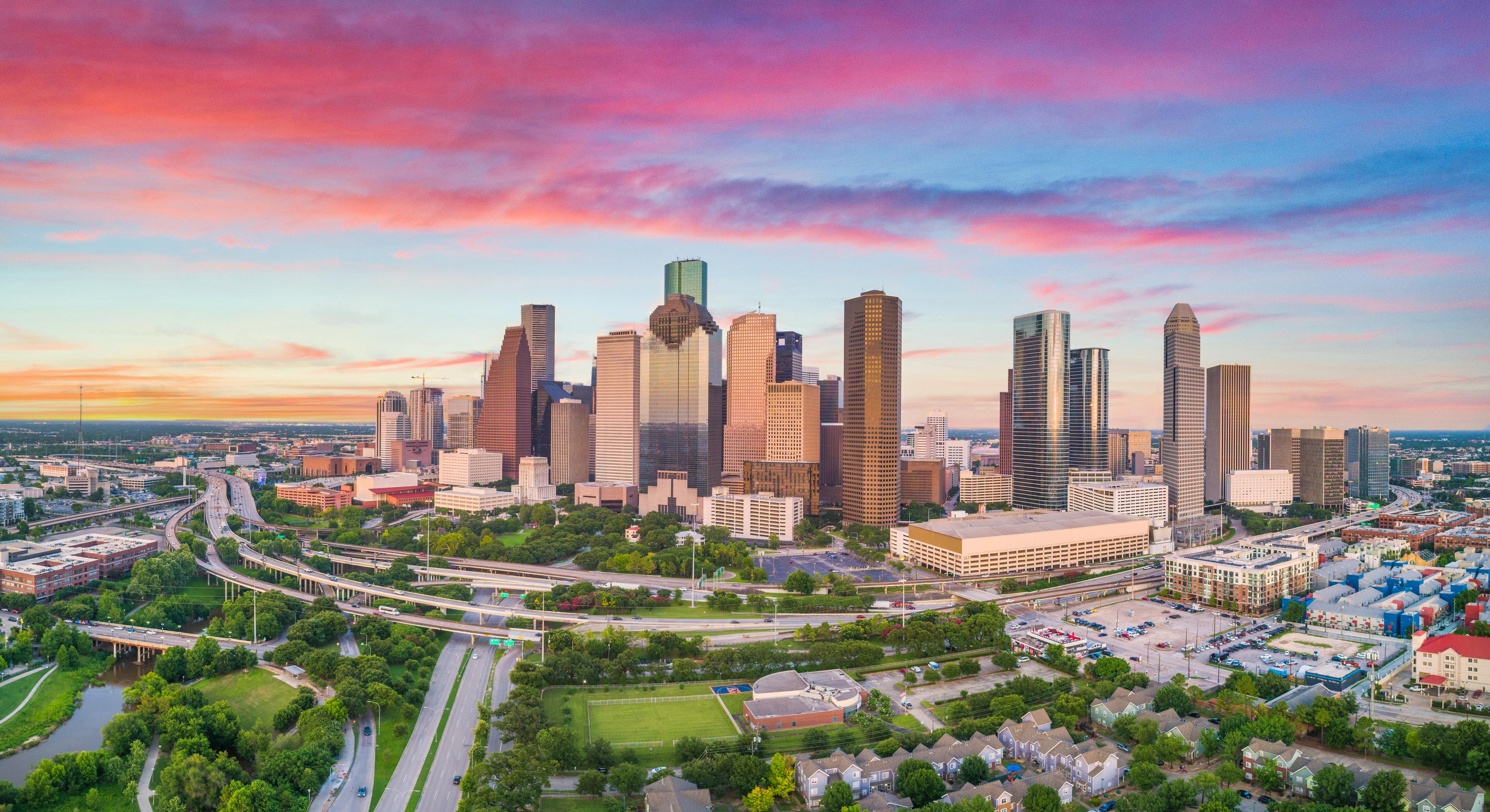 A panoramic aerial view of downtown Houston at sunset highlights the city's iconic skyline, dynamic highways, and lush green spaces under a colorful Texas sky.
