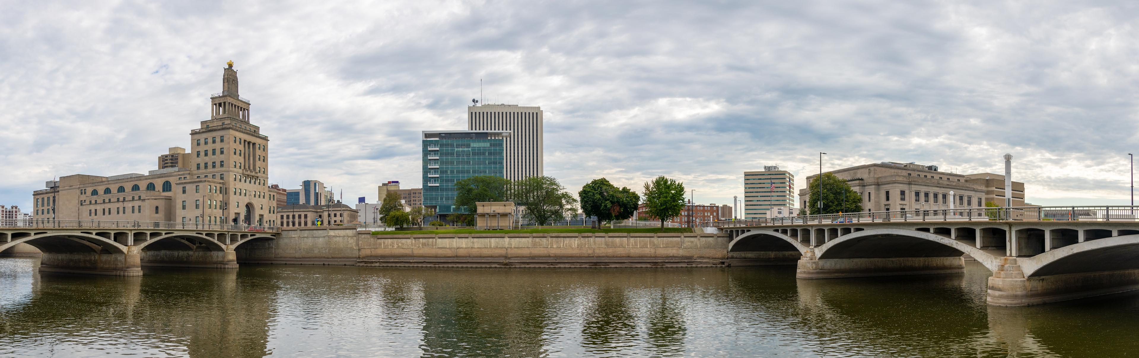 Downtown Cedar Rapids, Iowa, as seen from across the Cedar River, showcasing the City Hall, modern and historic buildings, and two bridges spanning the water. 