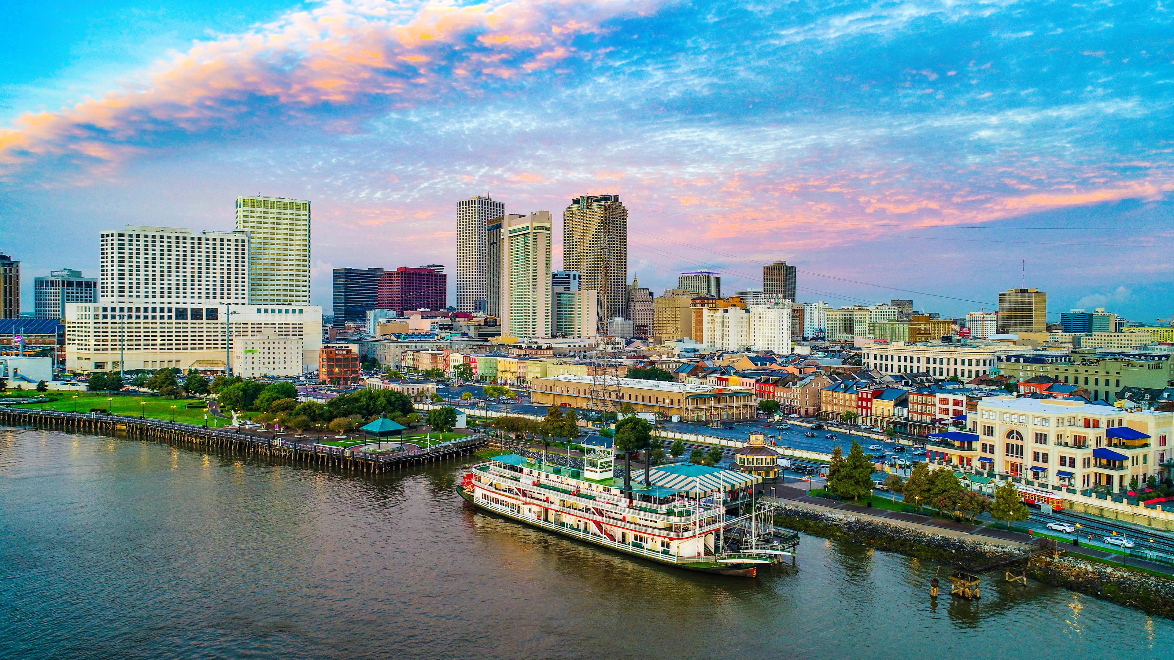 Vibrant aerial view of downtown New Orleans at sunset, with a paddlewheel steamboat docked along the Mississippi River and the city skyline glowing under a colorful sky.