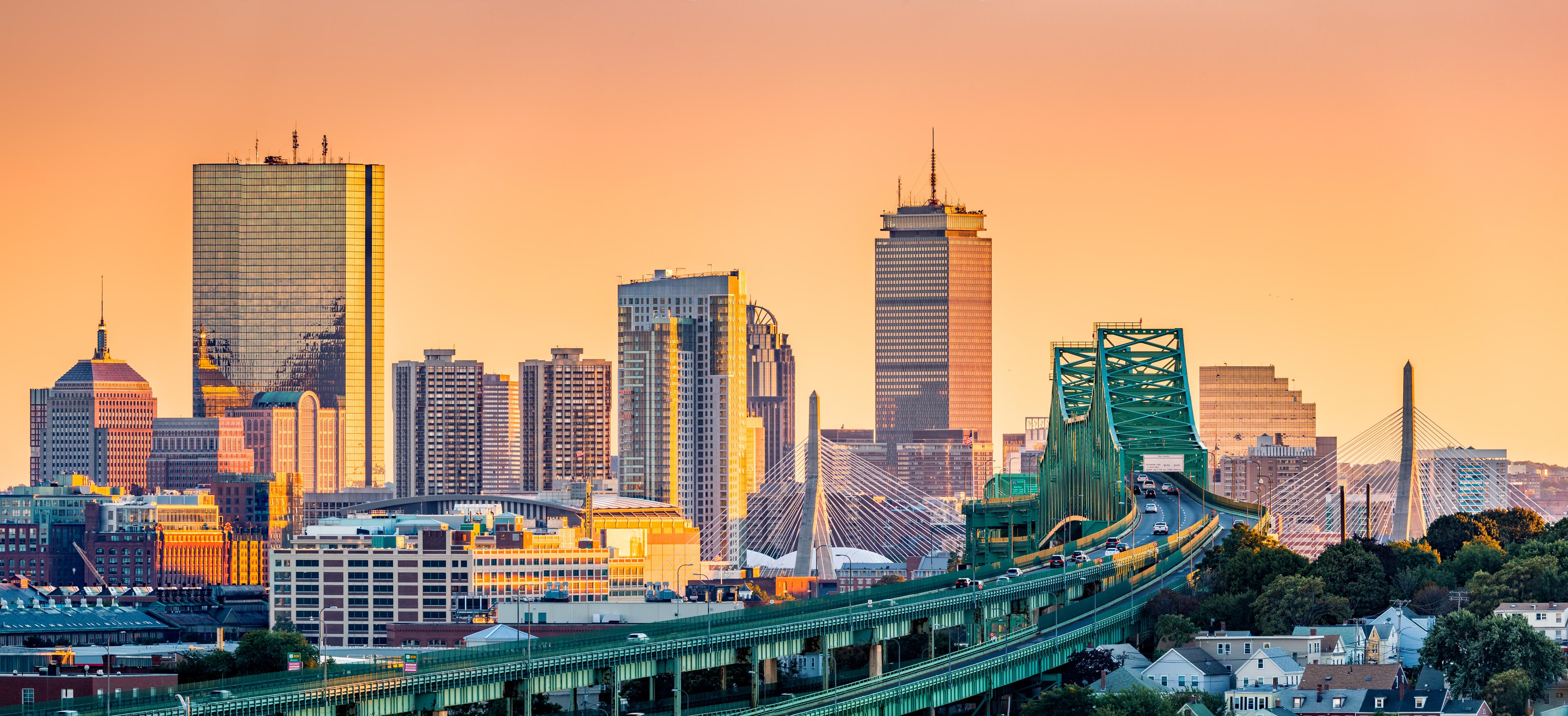 Boston skyline at sunset with the Zakim Bridge and Leonard P. Zakim Bunker Hill Memorial Bridge in the foreground, set against a warm orange sky.