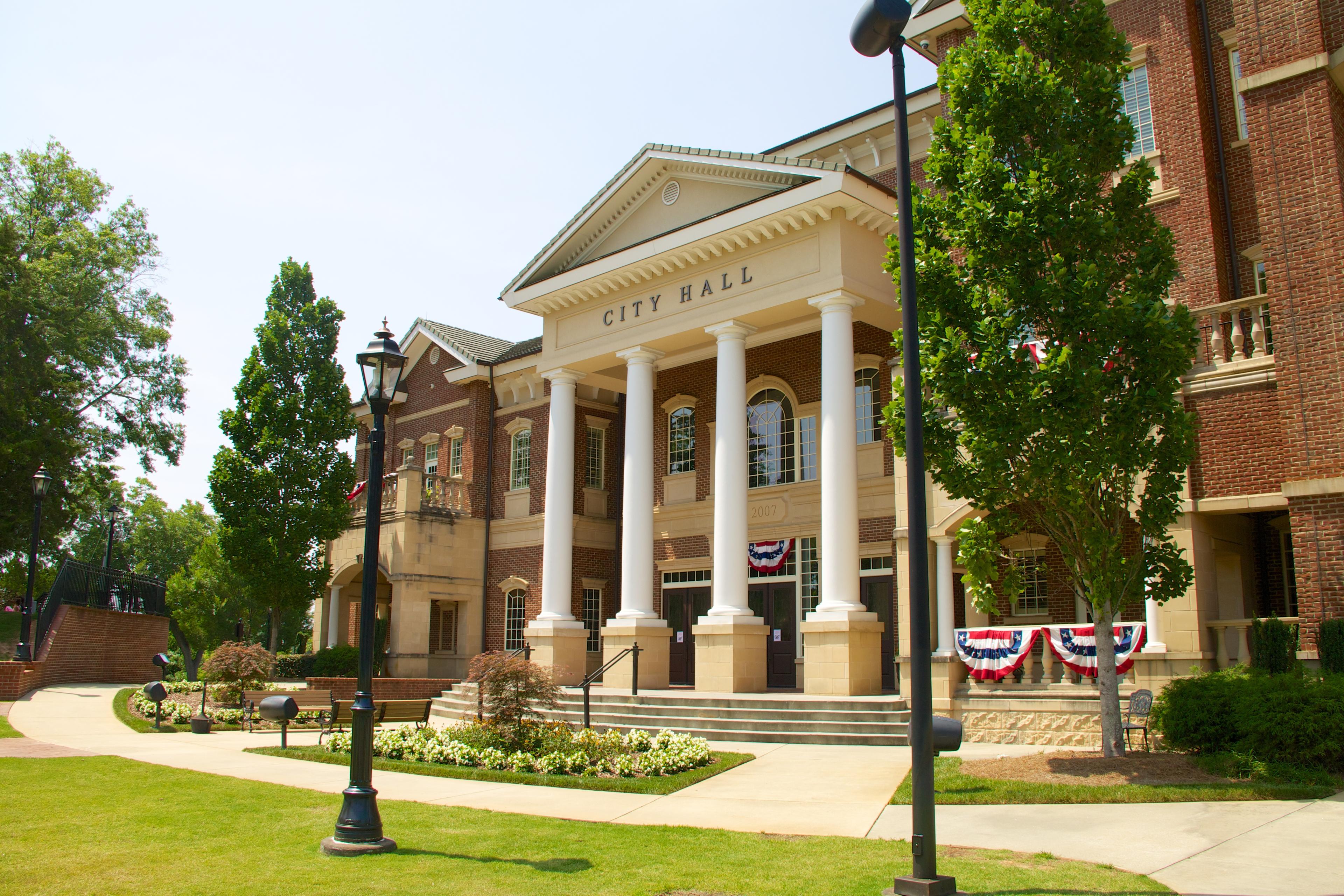 A brick city hall building with white columns, surrounded by green trees, a manicured lawn, and decorative red, white, and blue bunting.